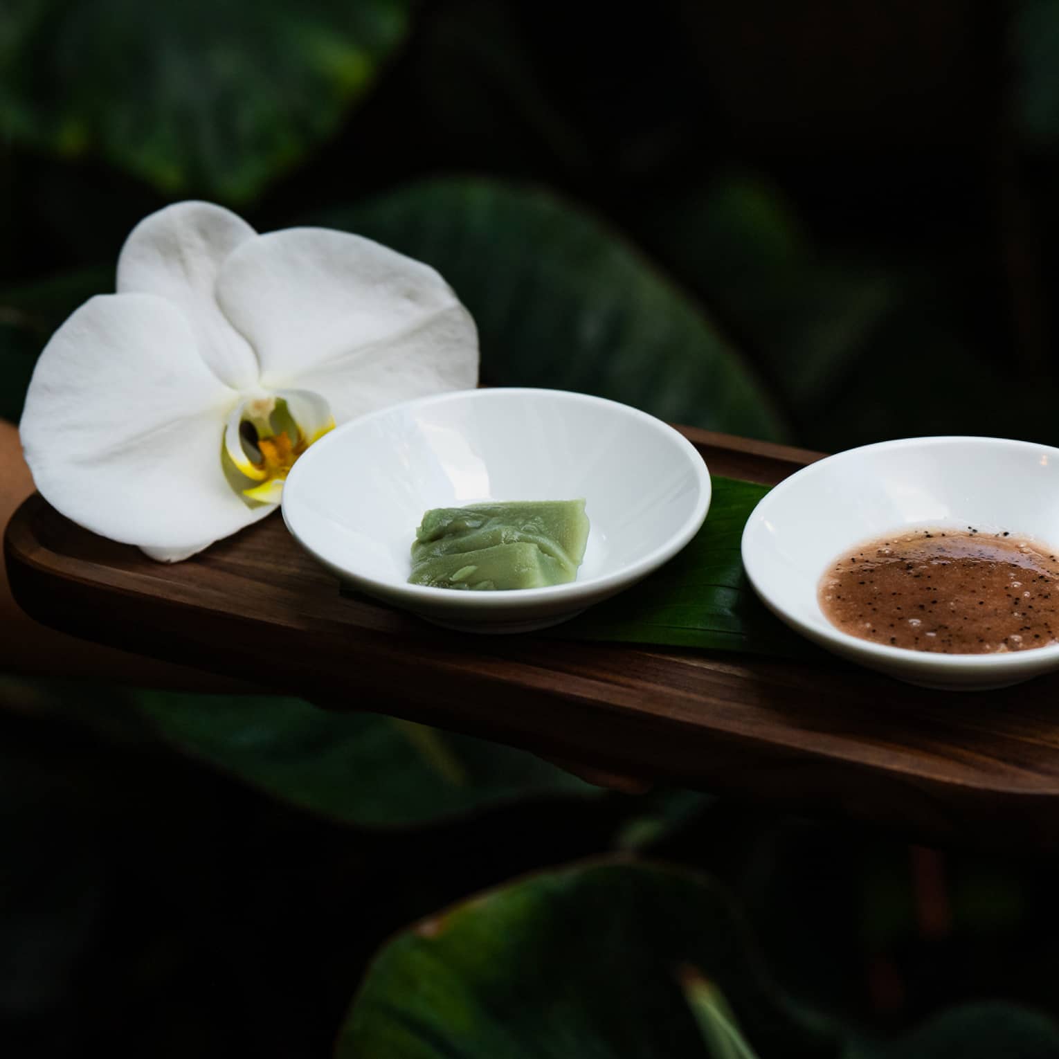 Rectangular wooden tray holds two small white bowls of spa ingredients and a large white flower