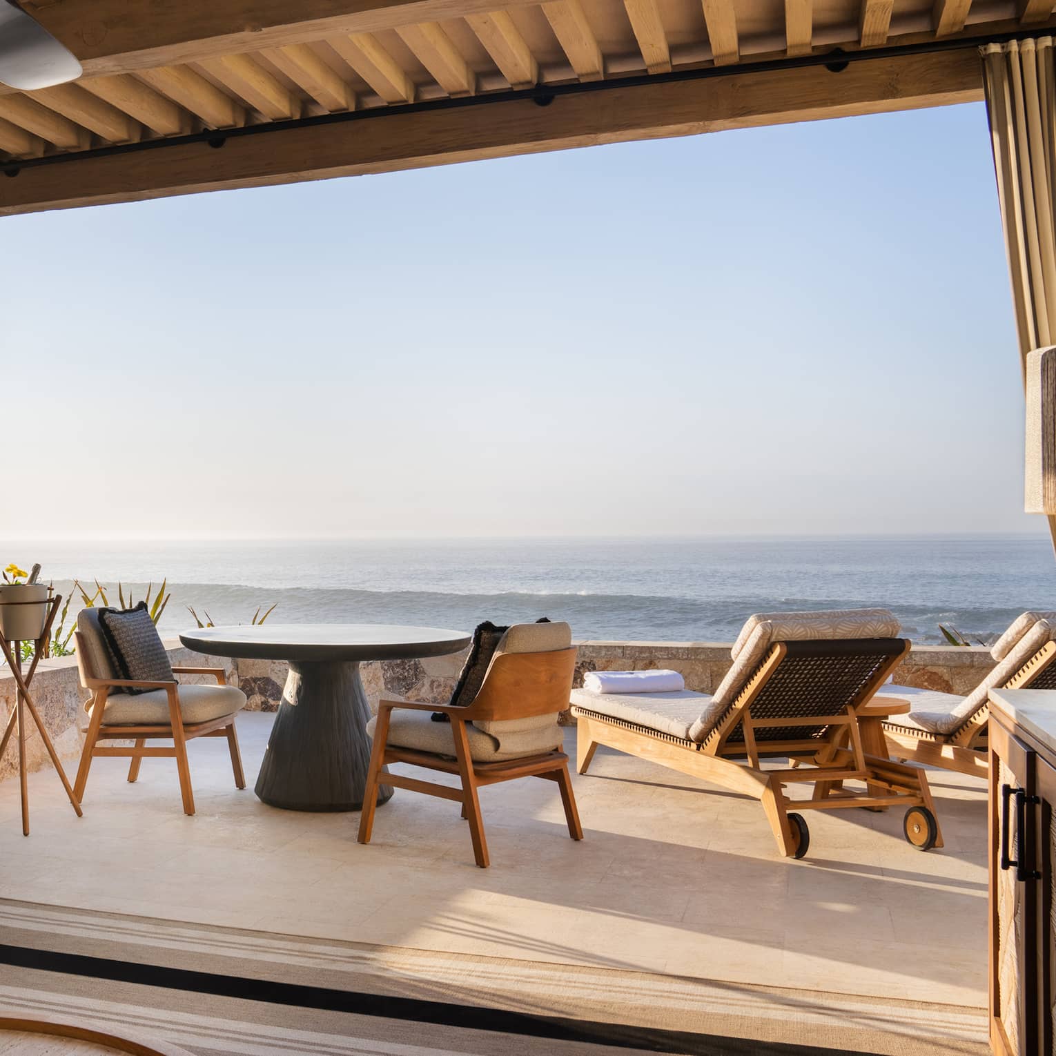 Oceanfront cabana with beige sofa, lamp and round wood coffee table under a roof with a round table and chairs and two lounge chairs out on the open-air patio overlooking the ocean