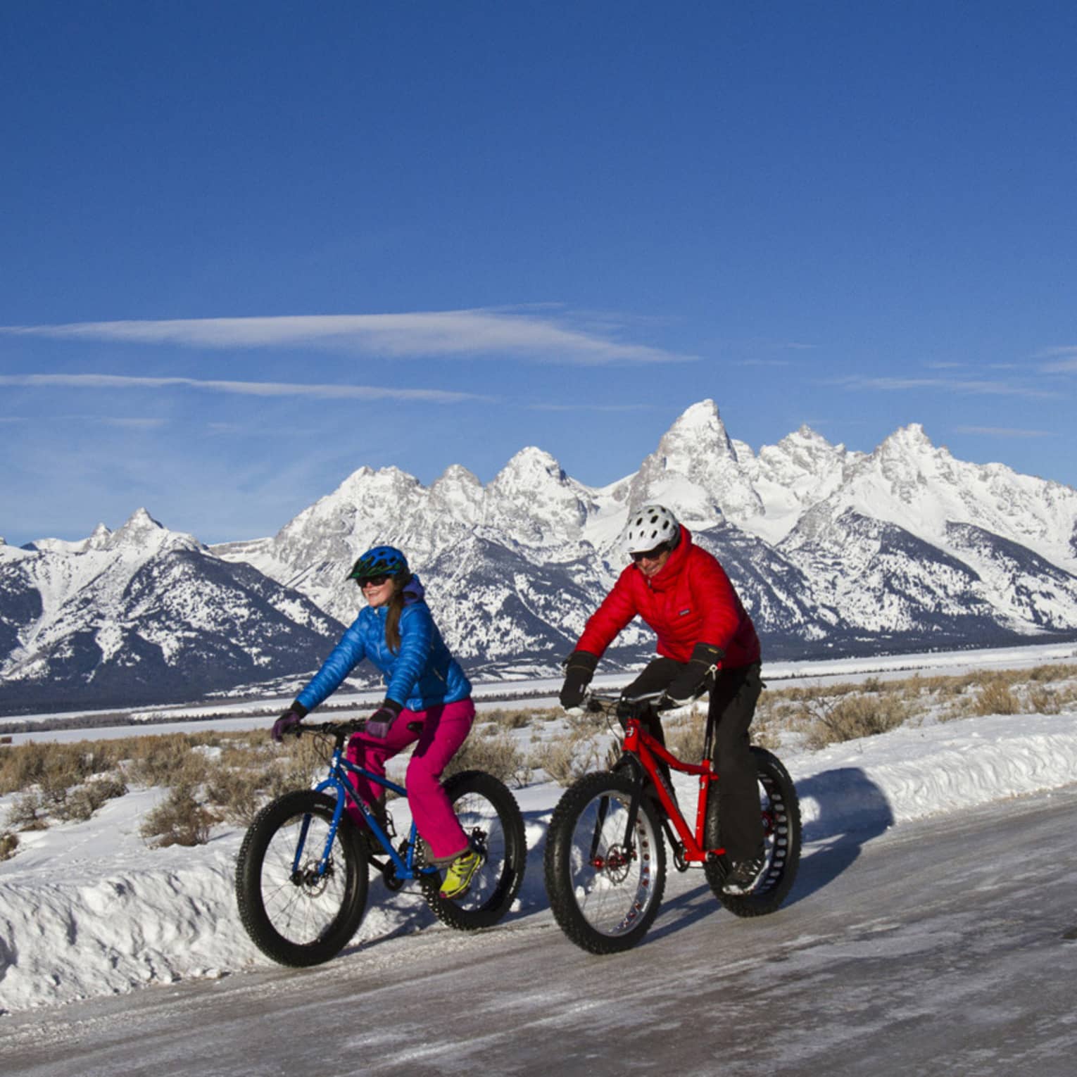 A man and woman bike down a path, snow-capped mountains and blue sky in the background