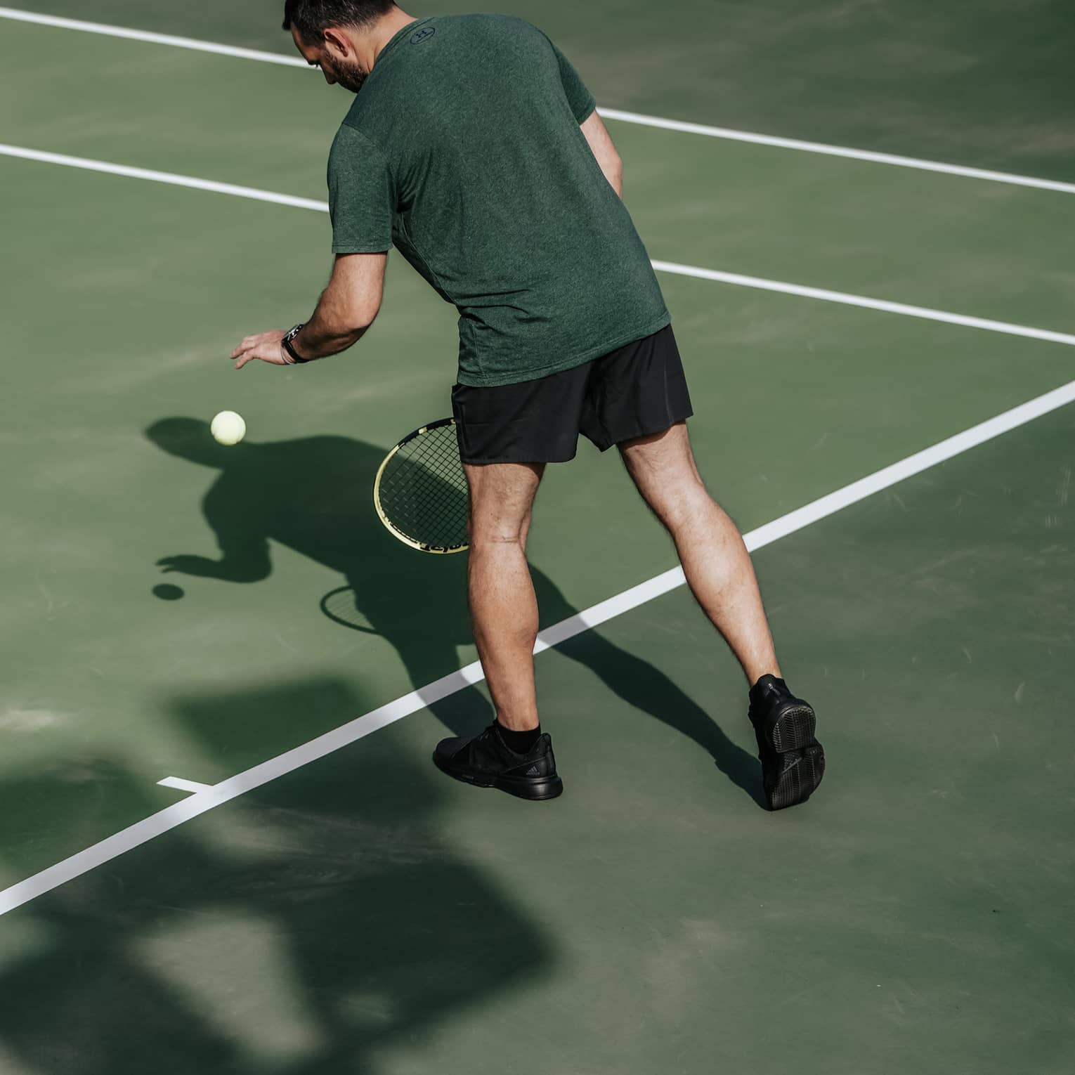 Man on tennis court bouncing tennis ball with left hand, preparing to serve