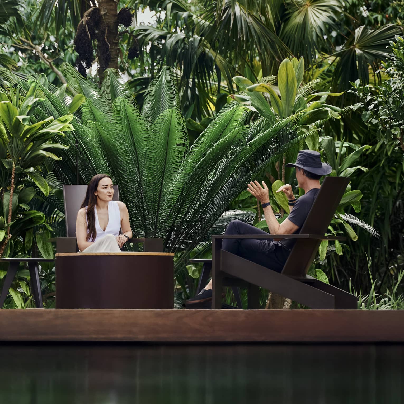 Surrounded by abundant tropical plants, two guests sit facing each atop a wooden platform over tranquil, glistening water.