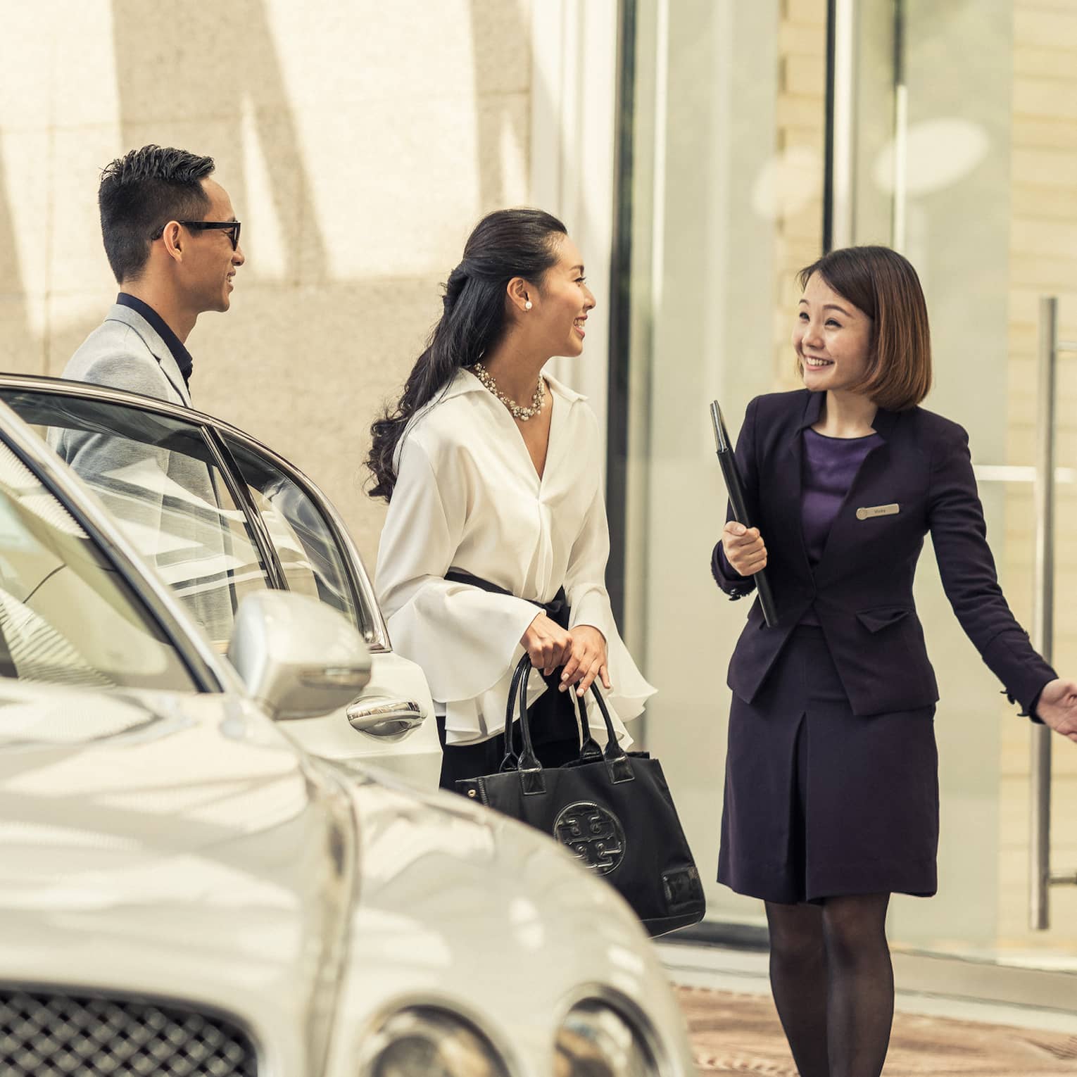 A person in a dark suit greets a couple near a luxury white car at the hotel entrance. The couple appears enthusiastic and engaged in conversation.
