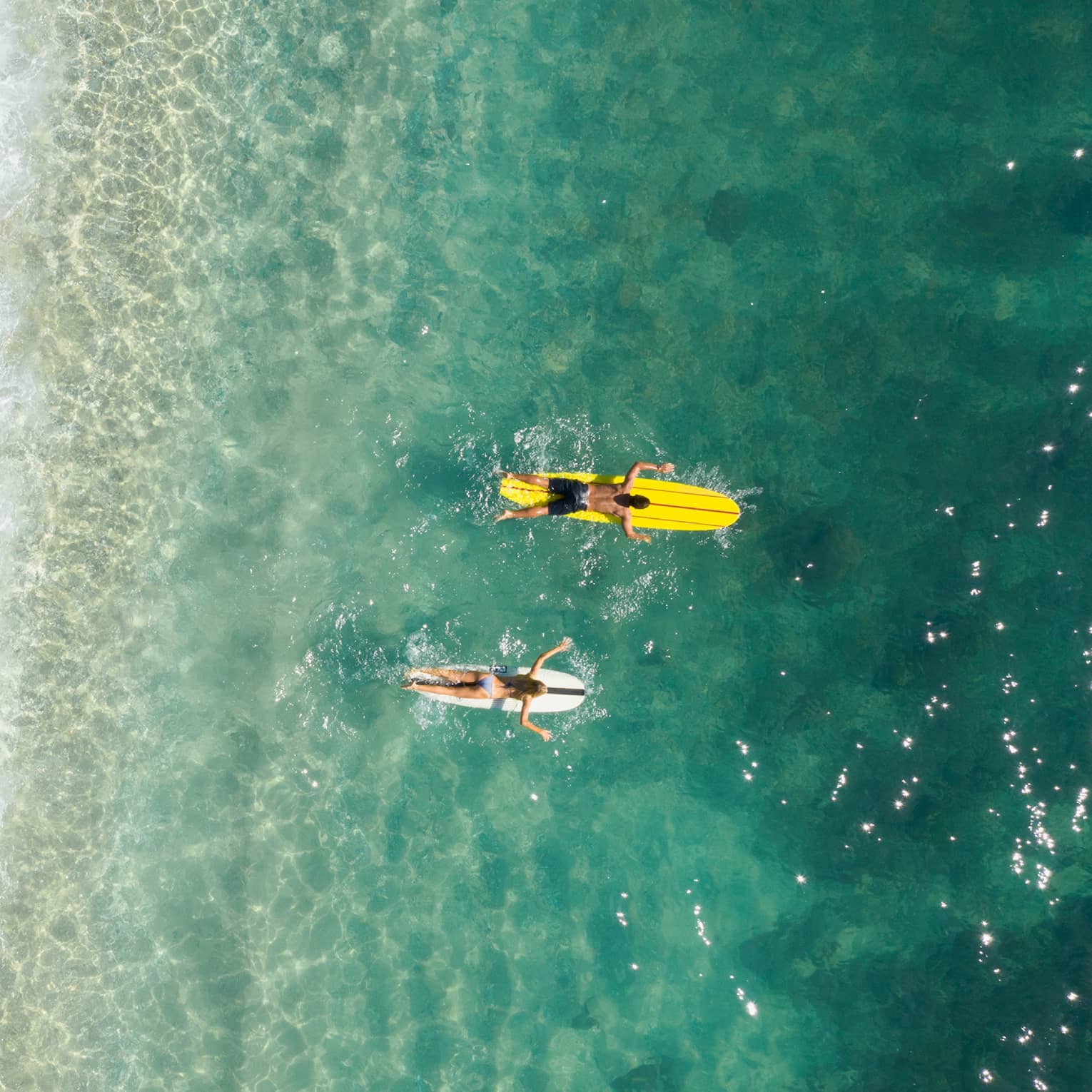 Bird’s-eye view of two people paddling their boards on their stomachs as frothy waves break on the beach behind them.