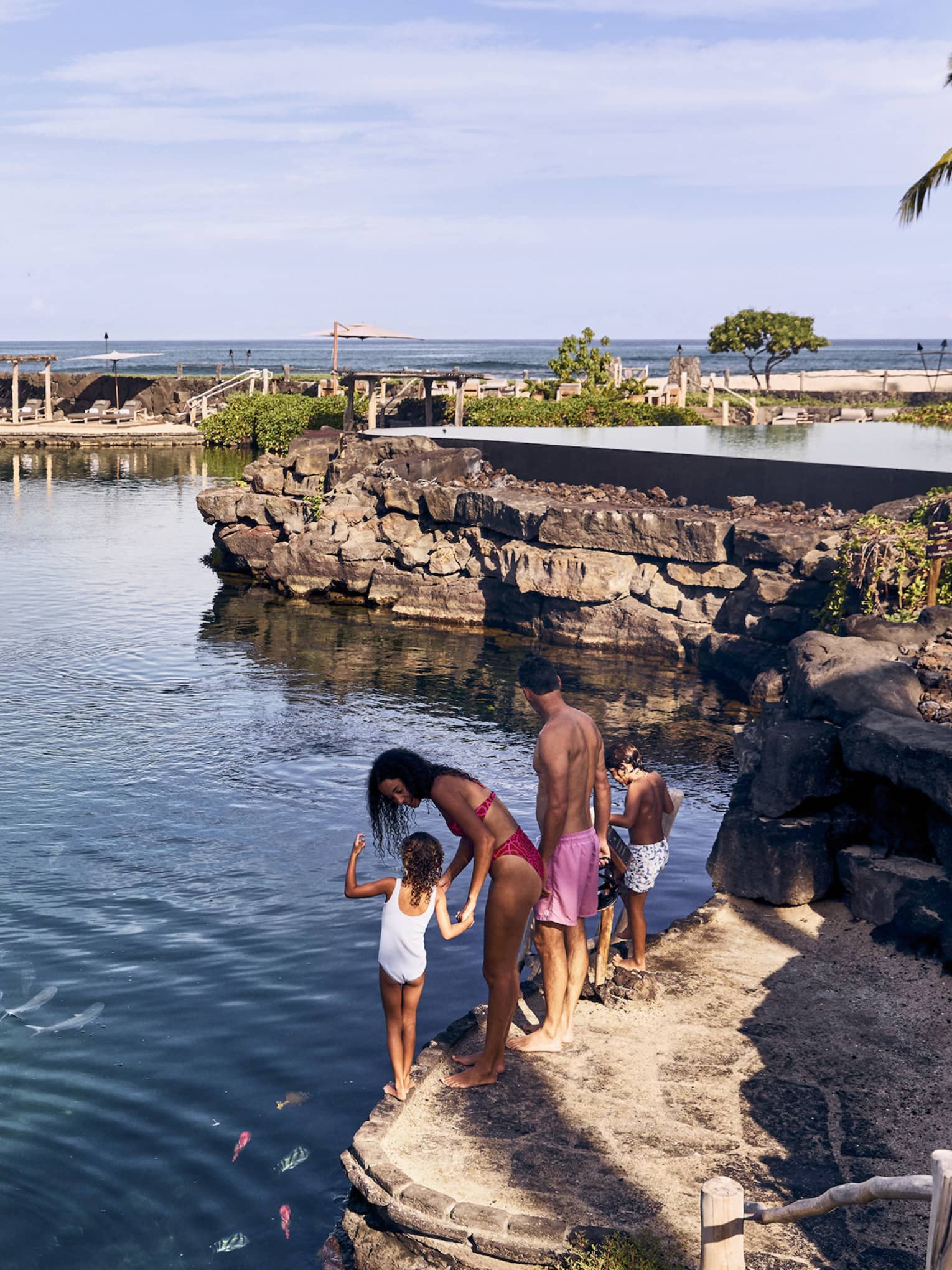 A family enjoying a natural swimming pond filled with fish with an infinity pool beside it and a beach in the background.