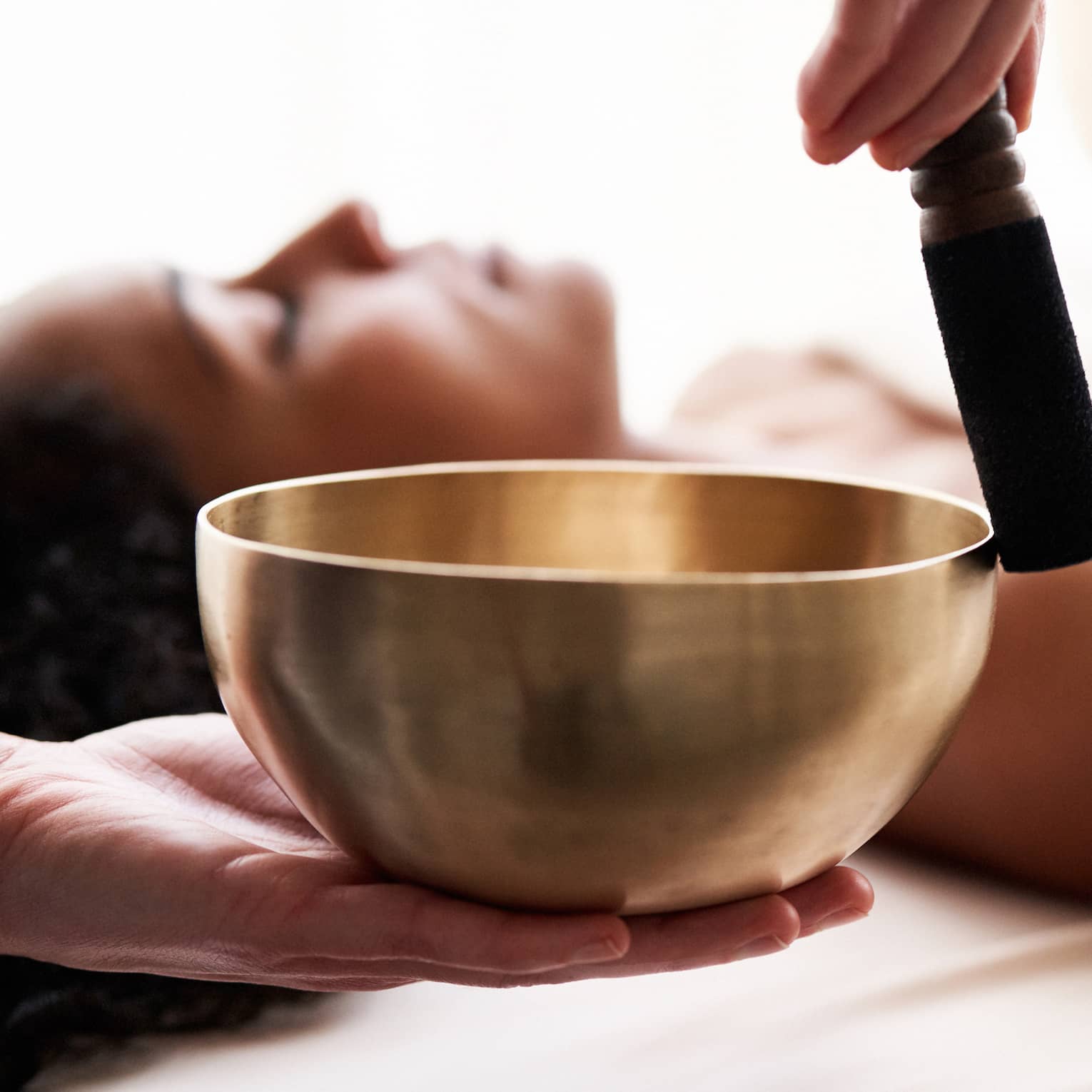 Close up of hands playing a singing bowl with a woman laying down on a spa treatment table in the background