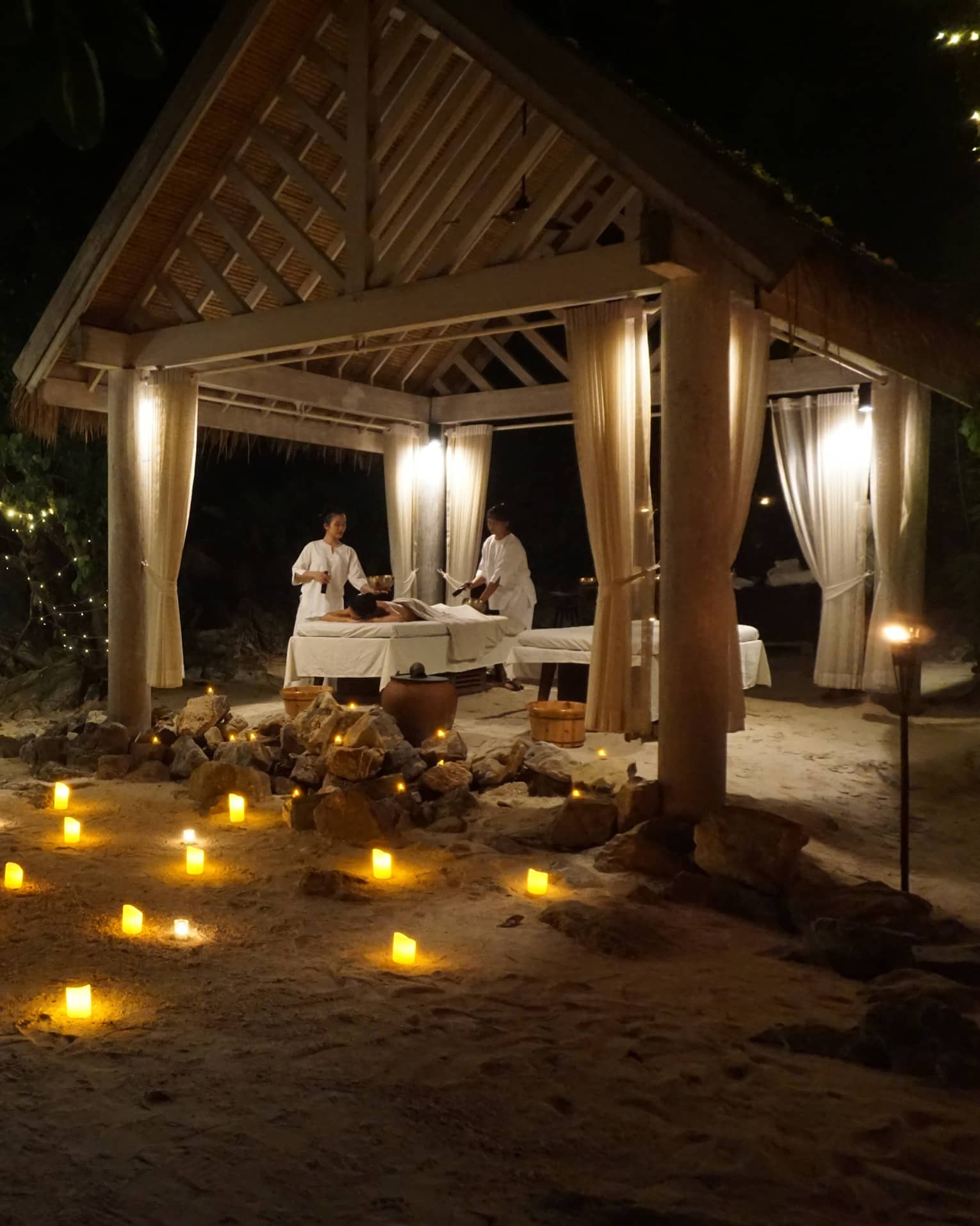 Spa treatment taking place under pergola in outdoor setting at night, with illuminated candles arranged on the ground