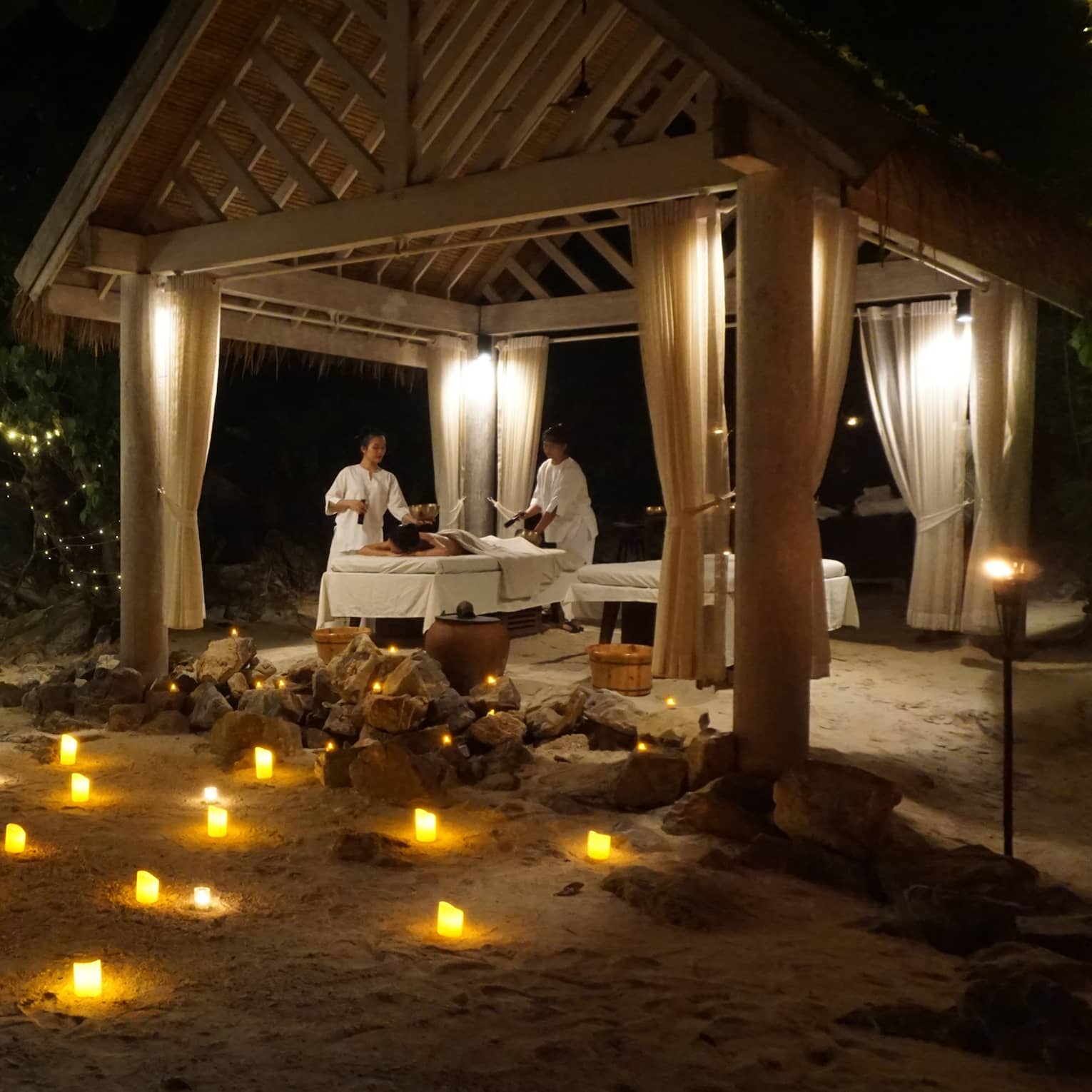 Spa treatment taking place under pergola in outdoor setting at night, with illuminated candles arranged on the ground