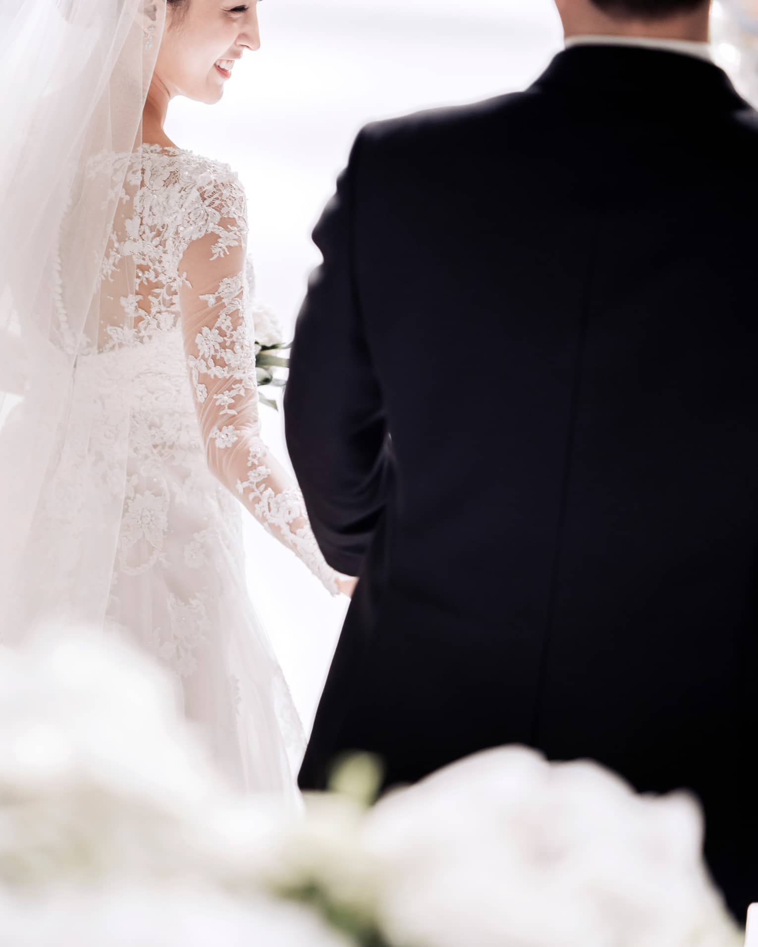 A bride in a white, lace, long sleeve gown smiles as she walks down the aisle with her groom. They are surrounded by artfully arranged white flowers.