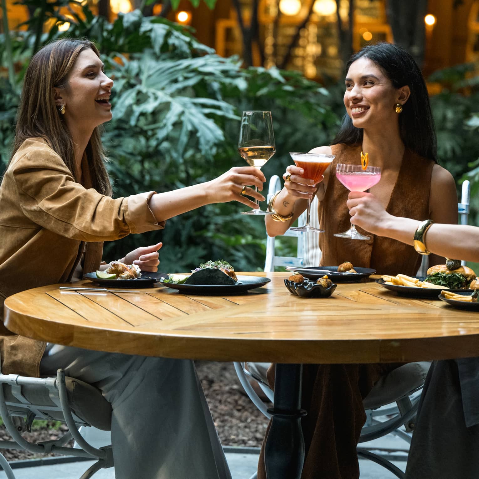 Three guests sit around a round wood table in an outdoor dining space and raise their glasses in a toast.