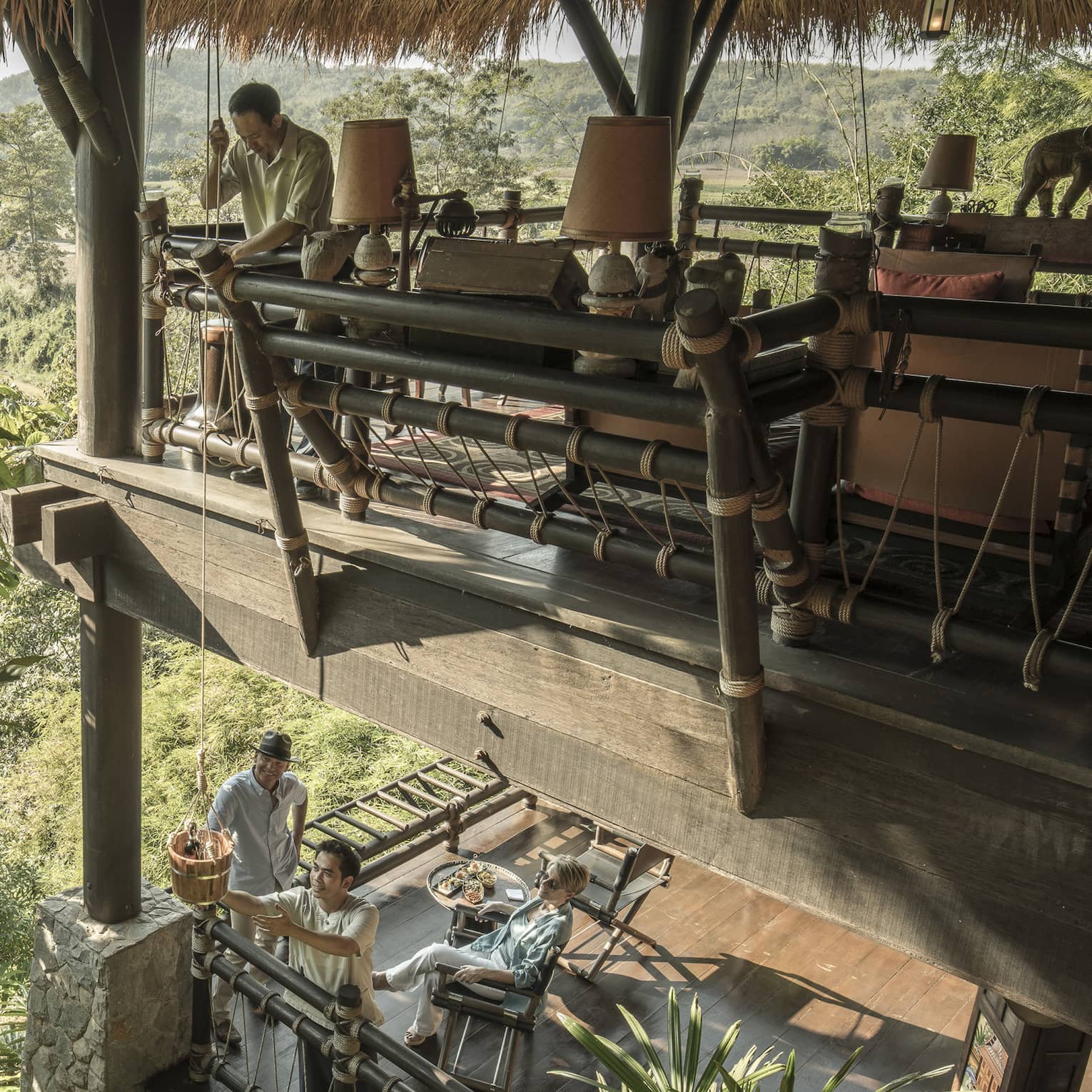People relaxing on the lower level of a two-storey open-air wooden deck as person passes basket of snacks down on rope