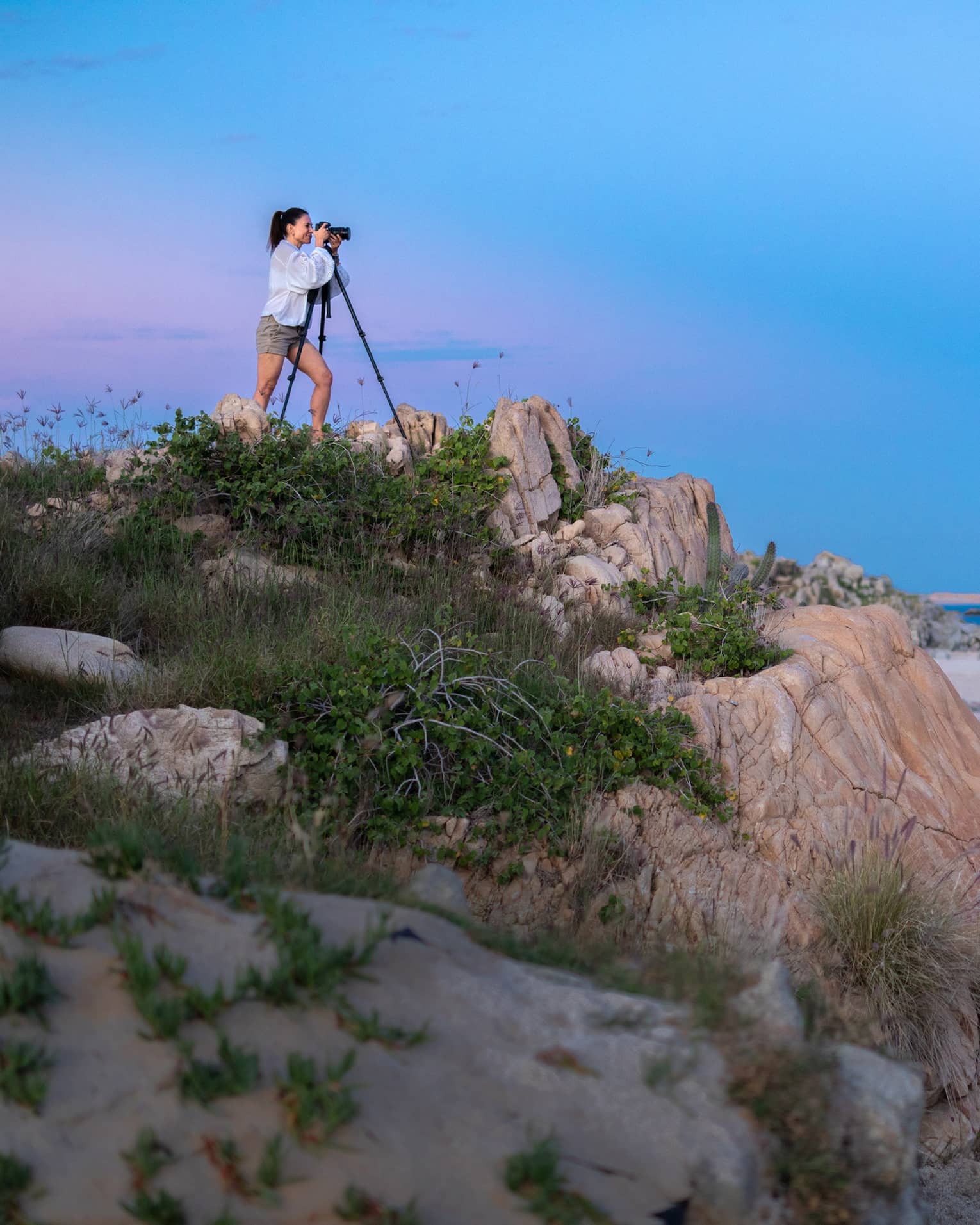 A photographer squints into a camera set on a tripod on a grassy rock outcrop overlooking calm, dark water under a full moon.