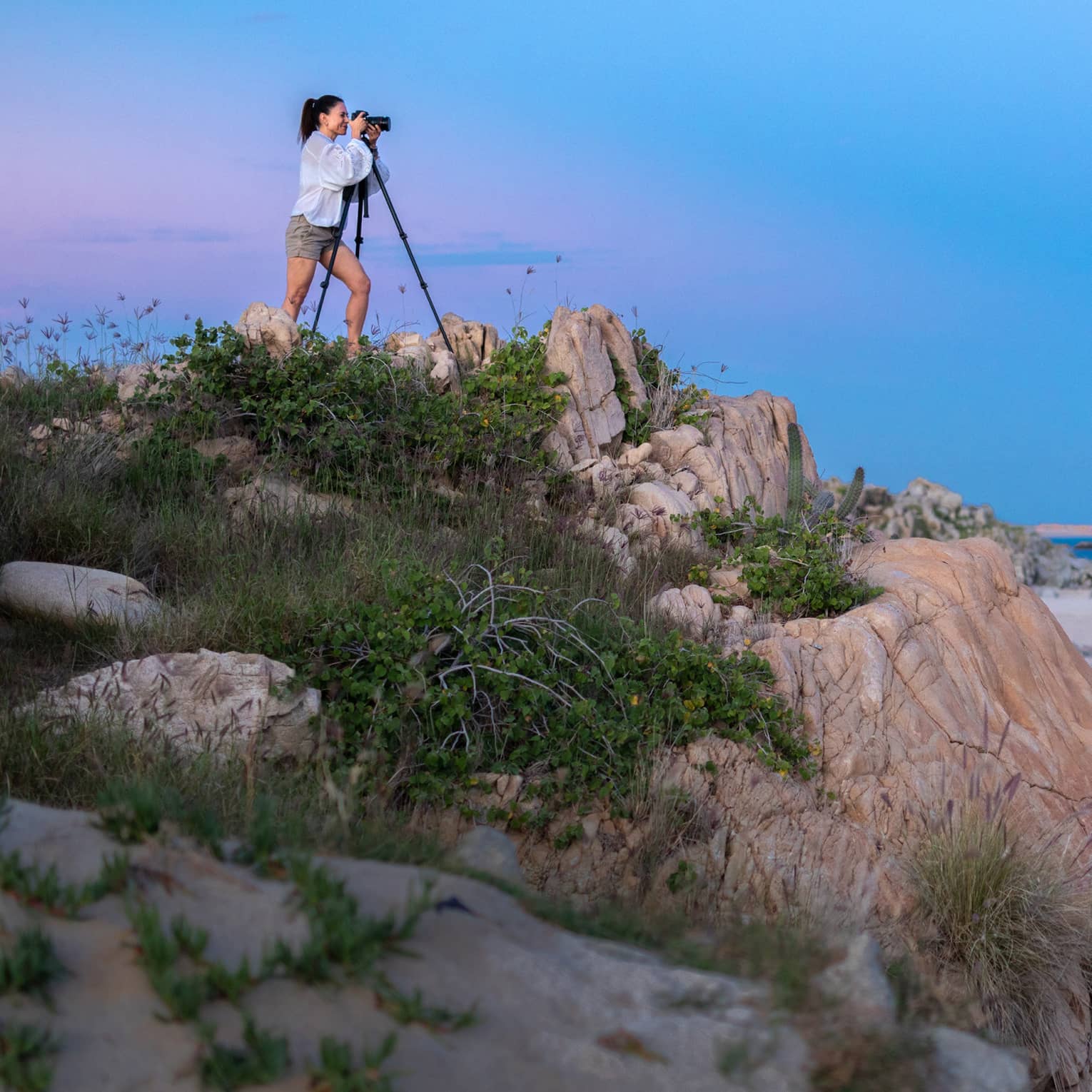 A photographer squints into a camera set on a tripod on a grassy rock outcrop overlooking calm, dark water under a full moon.