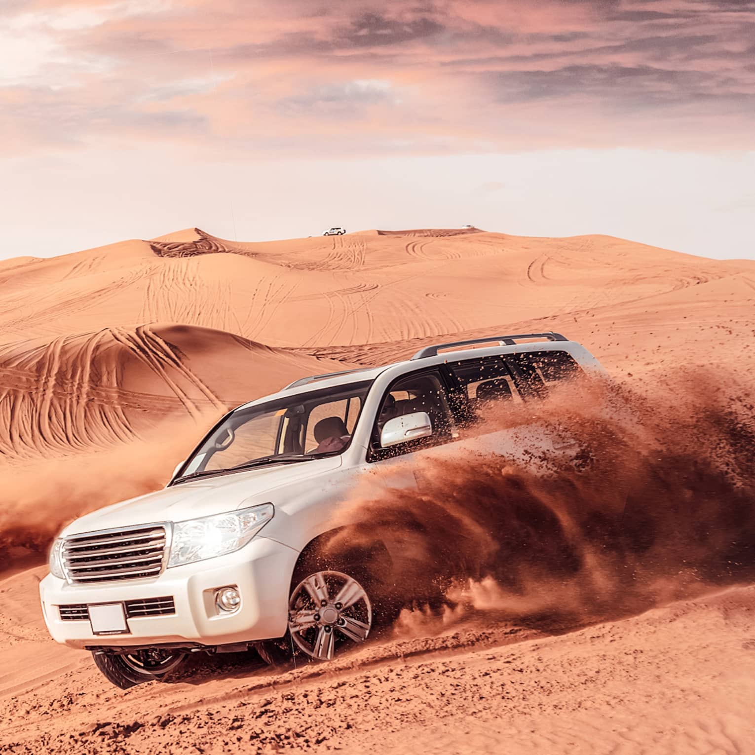 Sand flies from tires of two white SUV's driving across dunes
