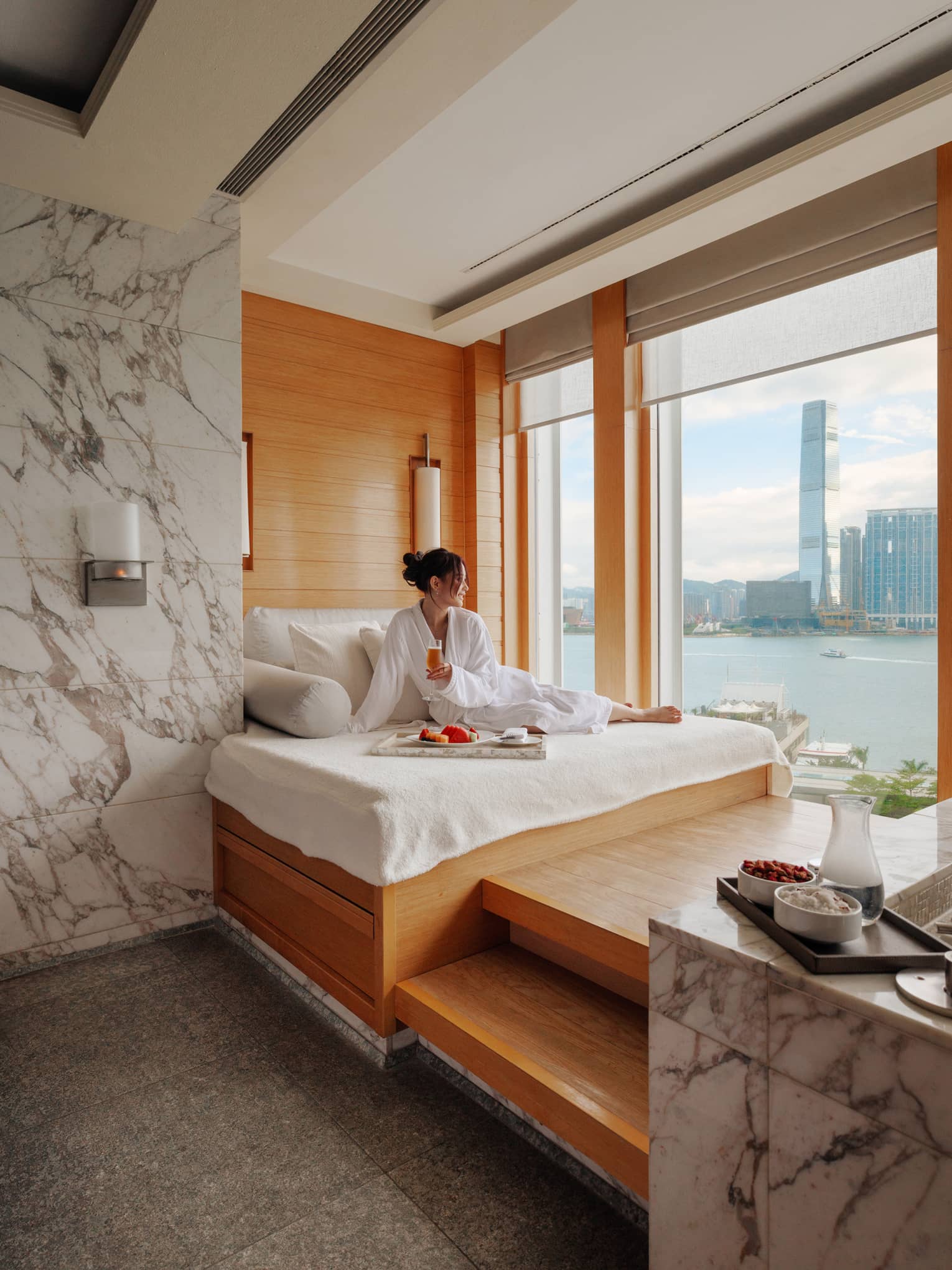 Woman in spa bathrobe sits on windowside chaise looking out to Victoria Harbour at Four Seasons Hotel Hong Kong