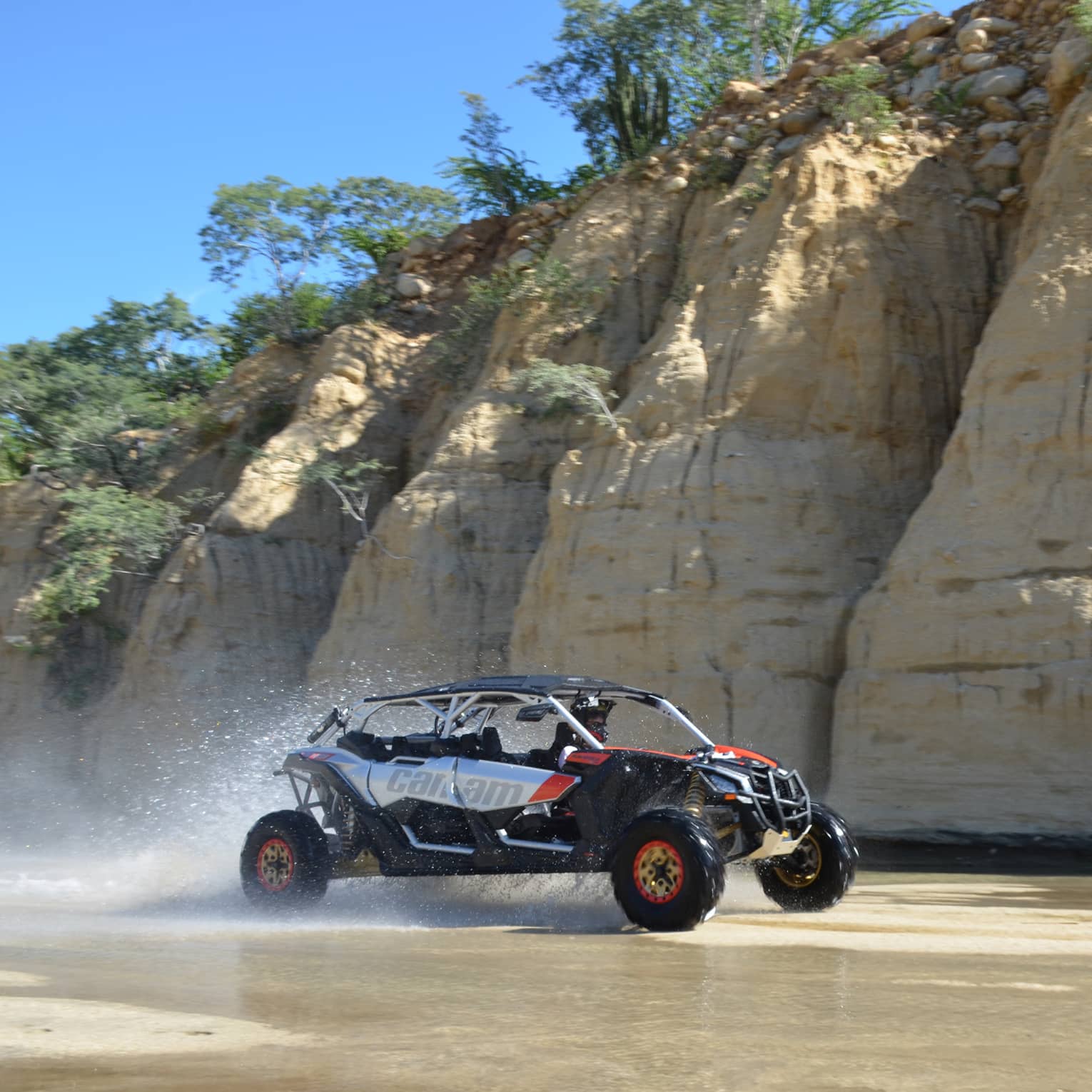 Water sprays off the back of a sporty off-road vehicle racing past deep vertical fissures in a tall crag topped with trees.