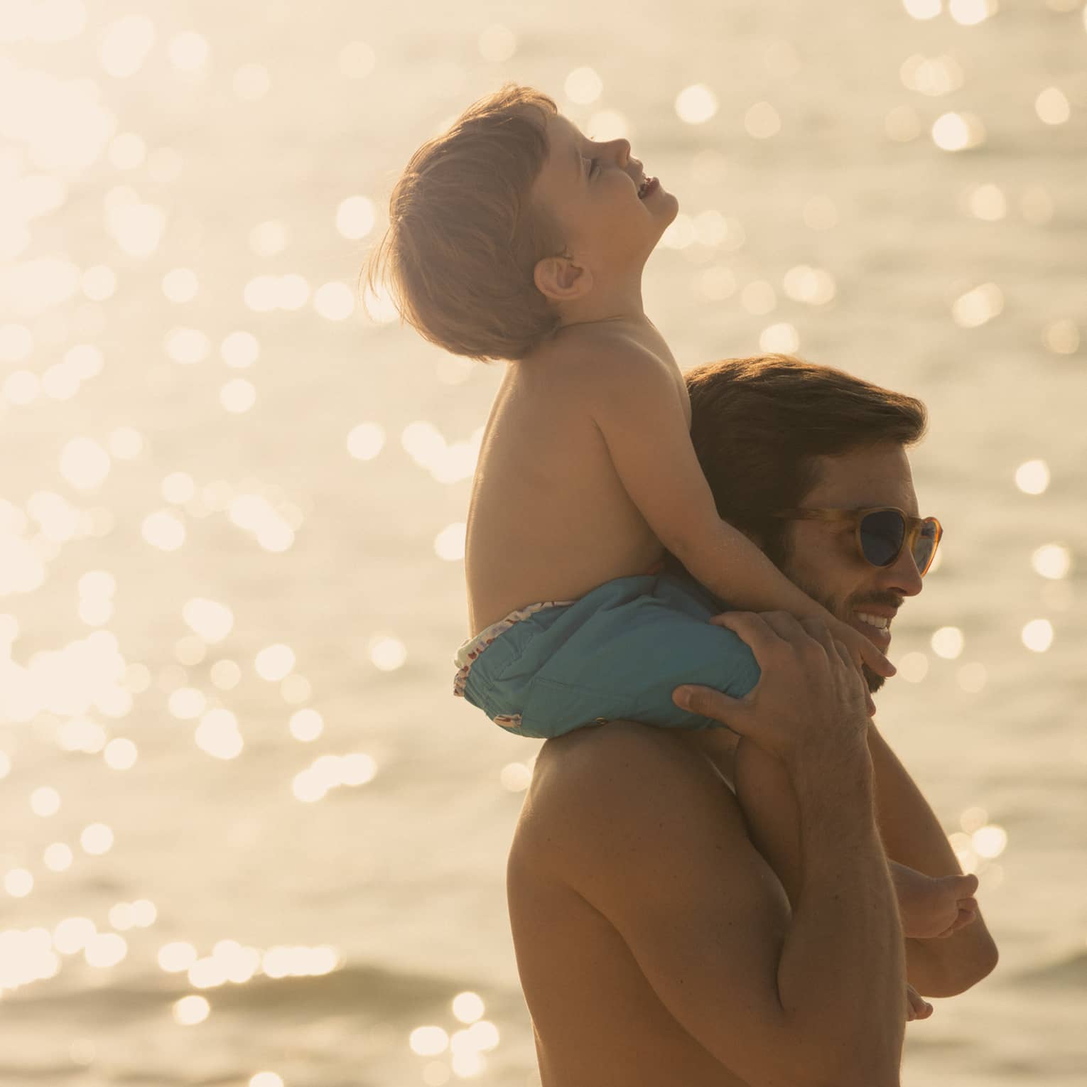 As the sun beams down over the glistening ocean water, a small child sits on their smiling parent’s shoulders and laughs.