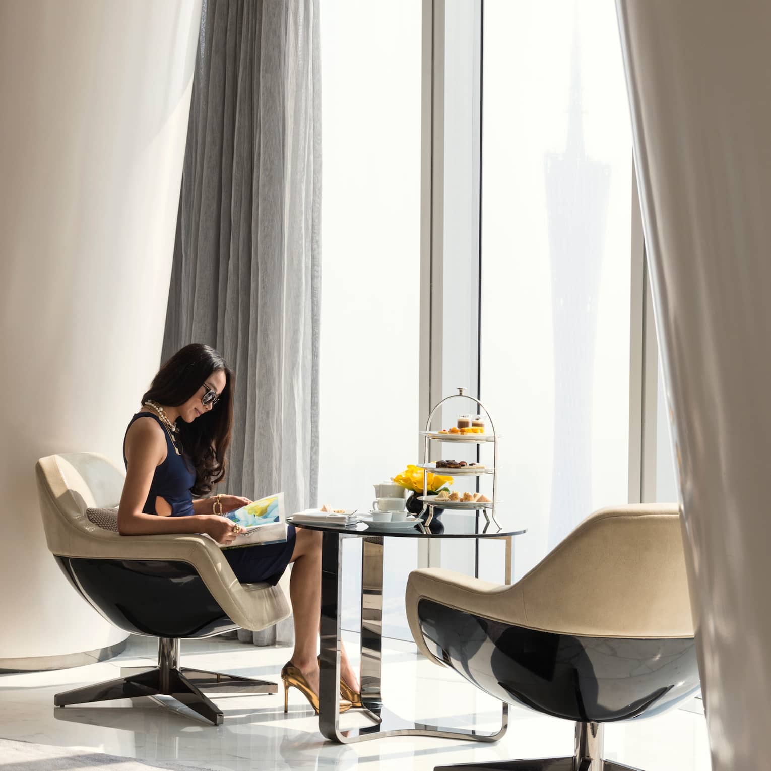 A guest sits in a neutral-toned room with a panoramic view and curved, modern design, a tiered tray of colourful fare nearby.