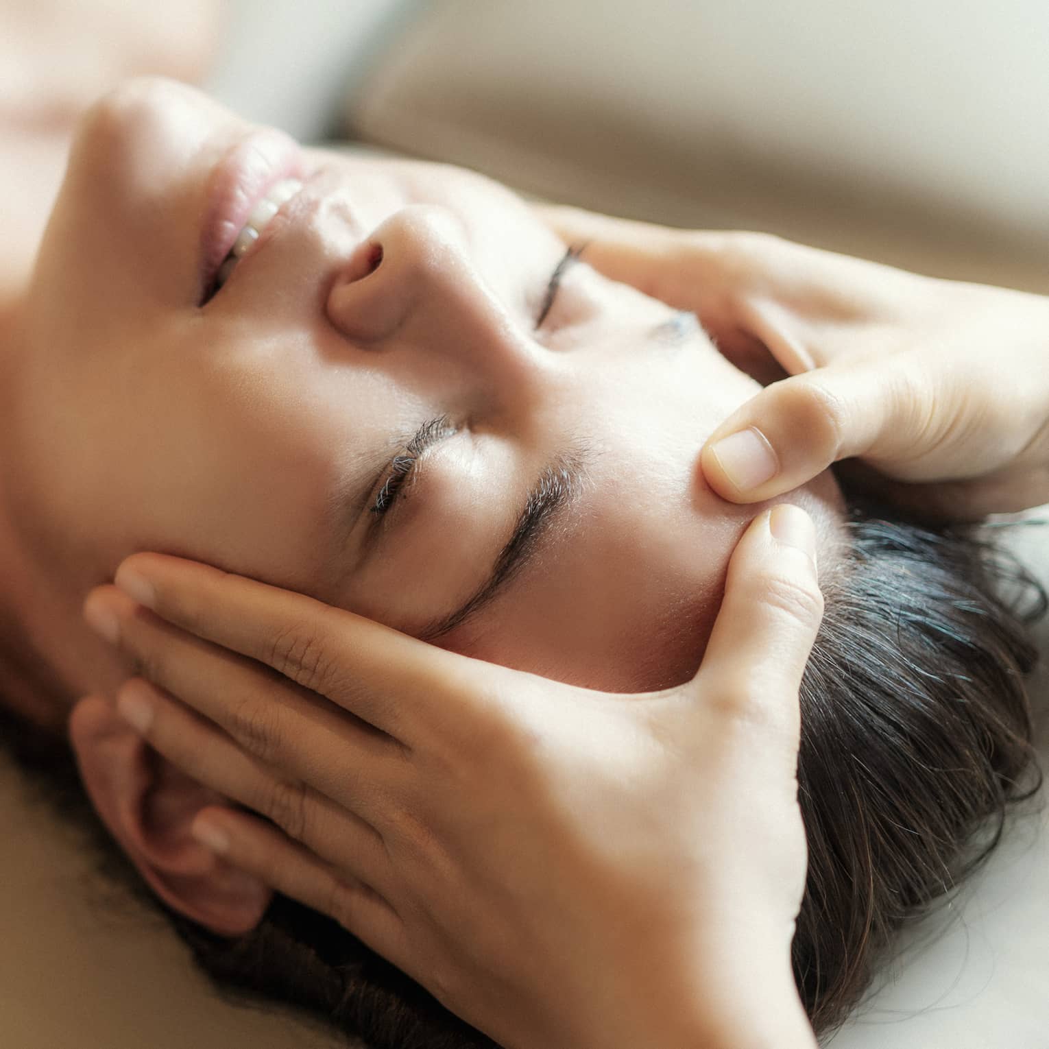 Spa facial, two hands rest on woman's forehead as she closes her eyes, lays under sheet on treatment table