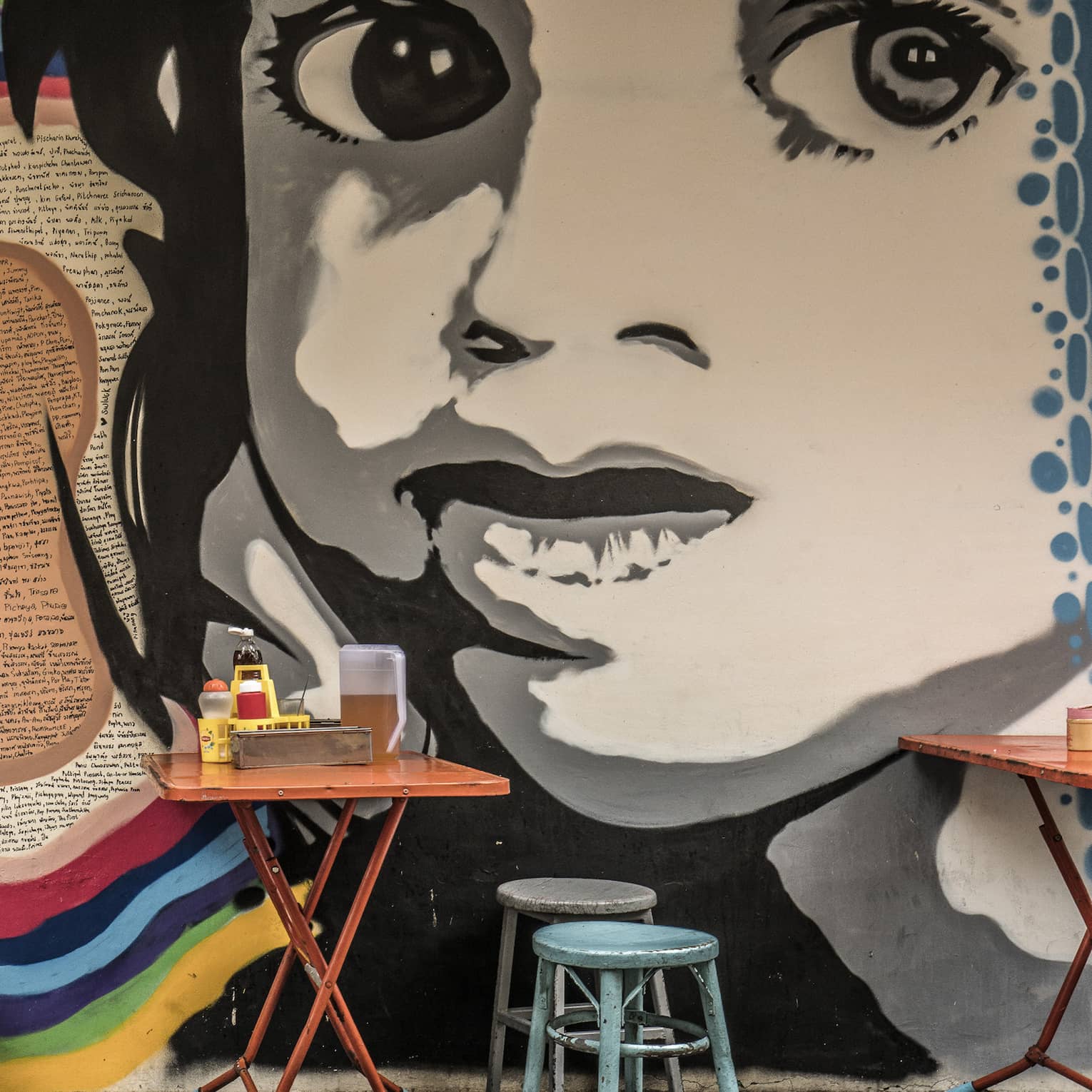 Street art mural featuring a large black-and-white face on a wall, with small, colourful dining tables and stools in front it