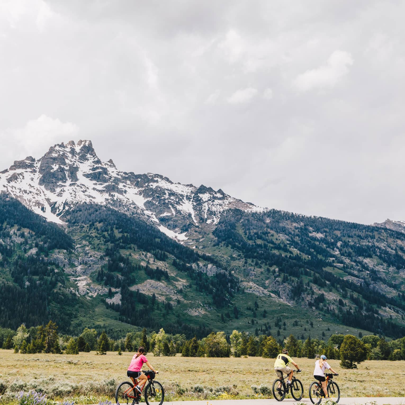 A man and two women cycle down a path on an overcast day with the mountains in the background