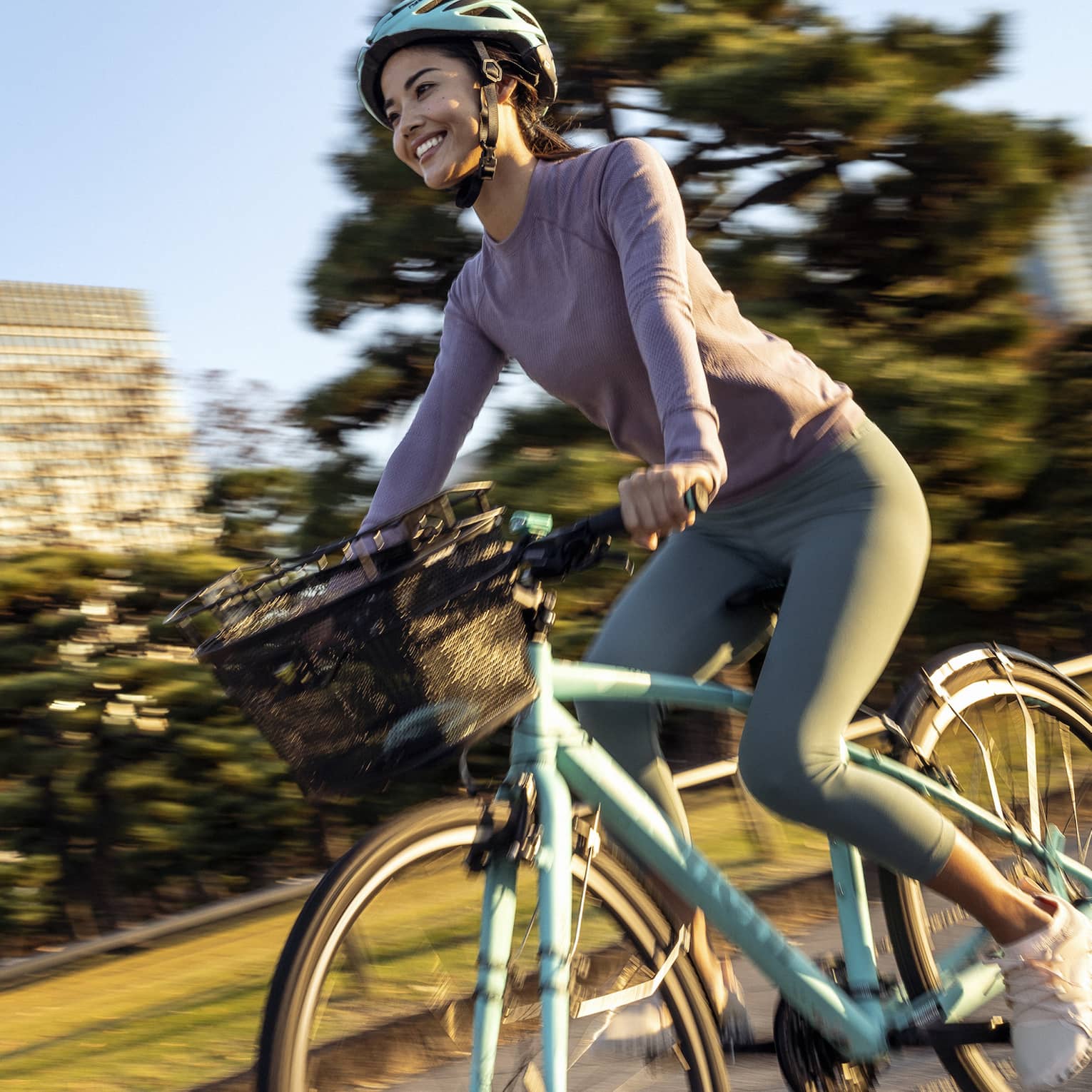 Smiling woman on teal cruiser bike with matching helmet rides by mature trees and skyscrapers of downtown Tokyo