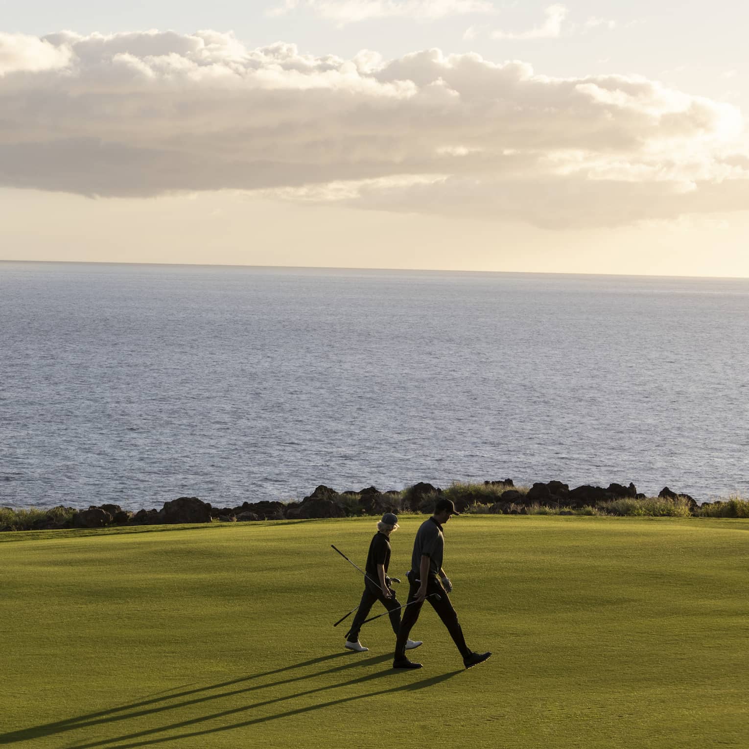 Two people with golf clubs, long shadows reaching out behind, walk a golf course set against calm ocean and fluffy clouds.
