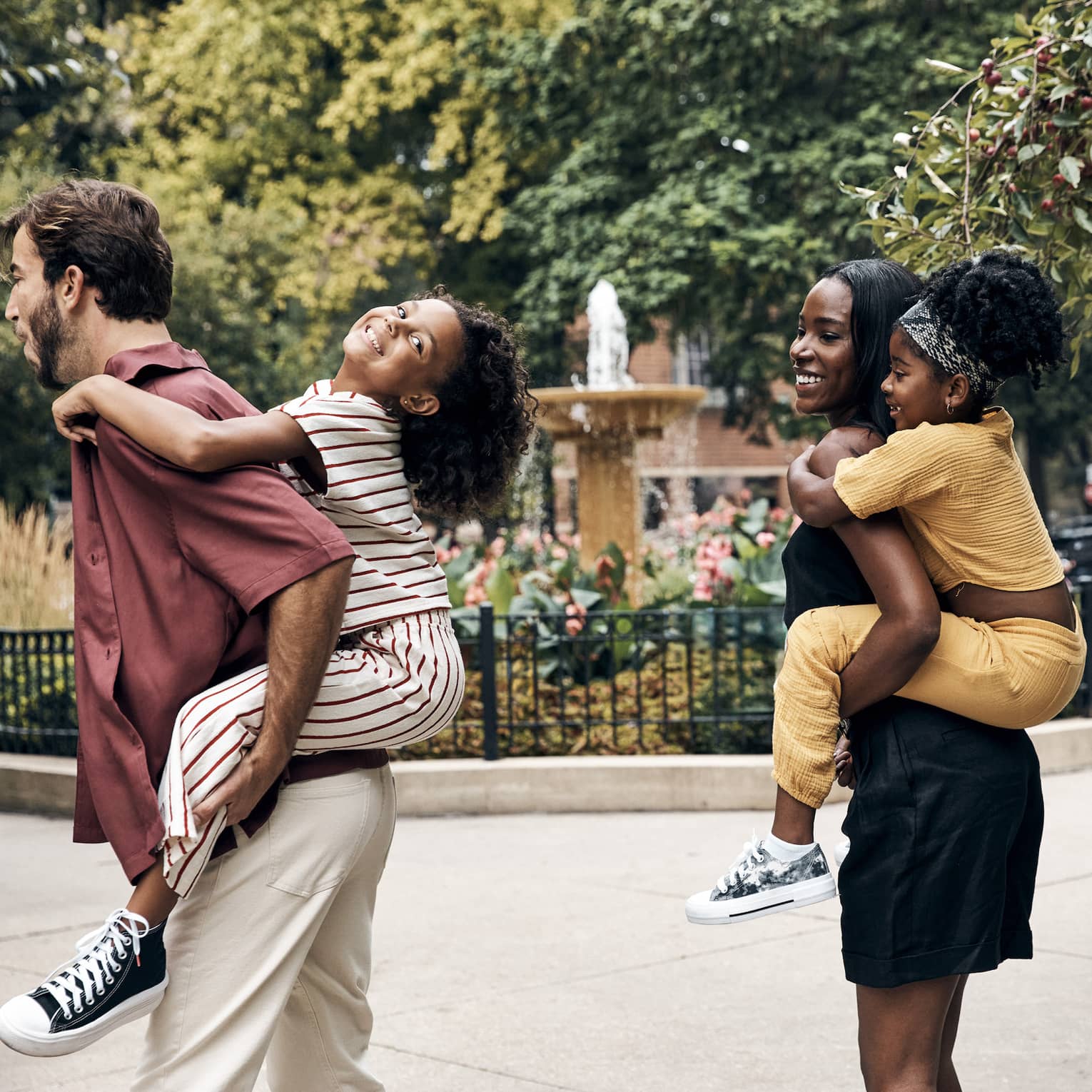 Two parents each holding a child on their back walk by a city fountain