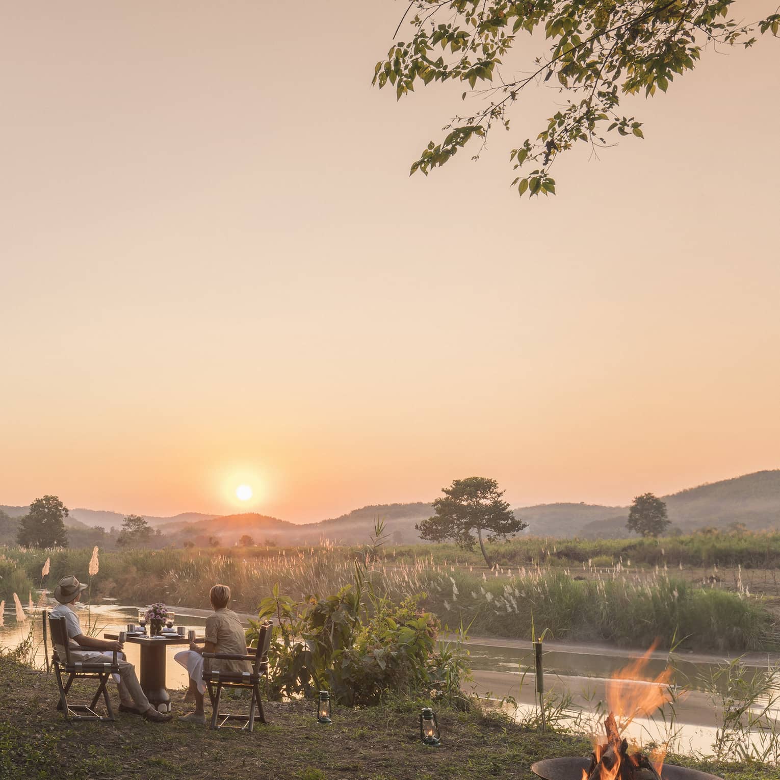Outdoor riverside dining setup at sunset with a campfire and mountain views