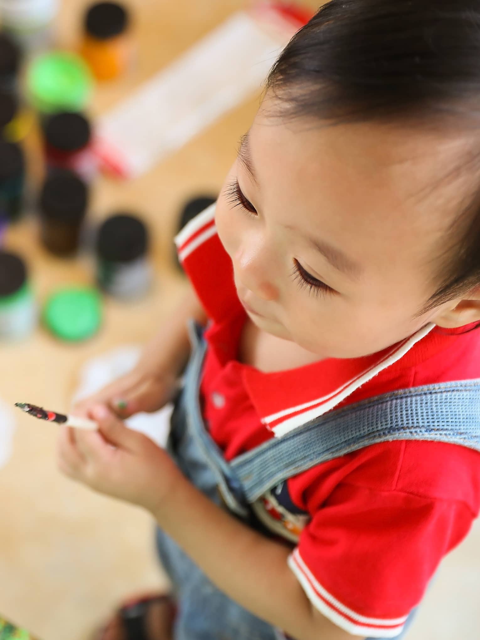 Aerial view of toddler boy standing in front of a canvas holding a paintbrush, a young girl sitting in the background surrounded by tins of paint