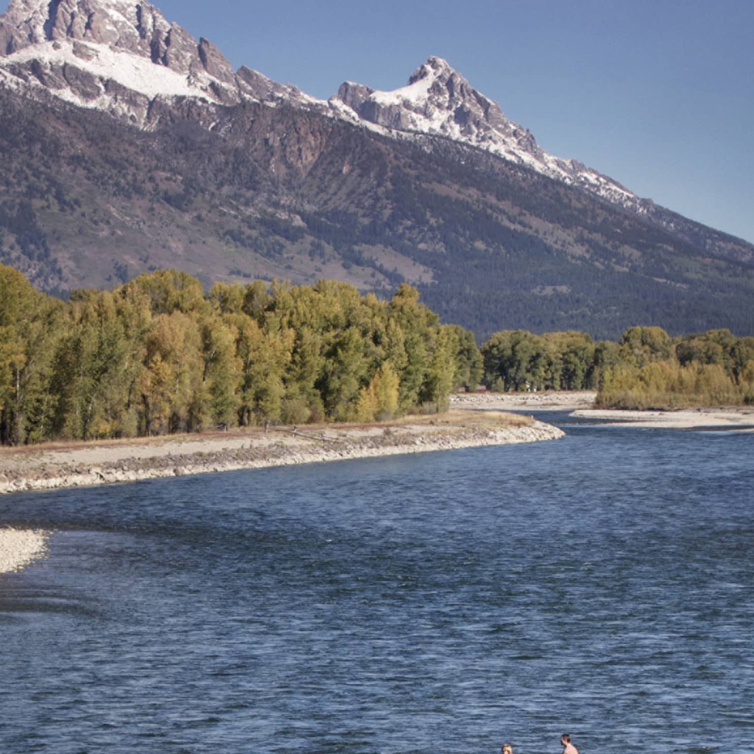 People fishing in boat on wide river by rocky shore, green trees, mountains