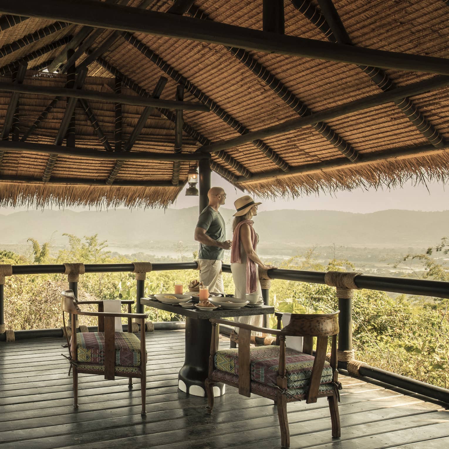 Couple enjoying mountain views from thatched wooden deck with vintage SUV parked below