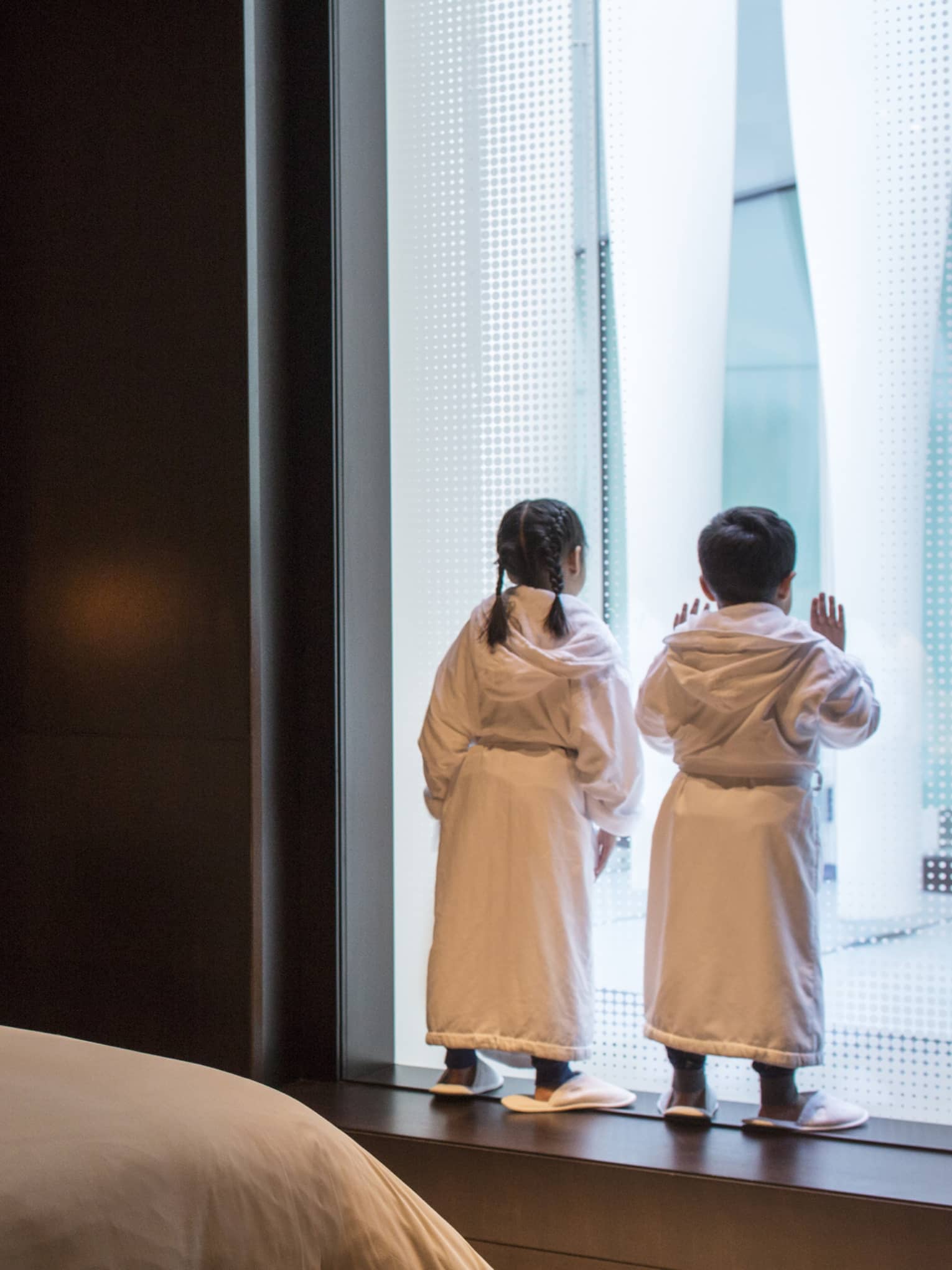 Young boy and girl in bath robes and slippers peer out the window at the city below, two teddy bears on the bed behind them