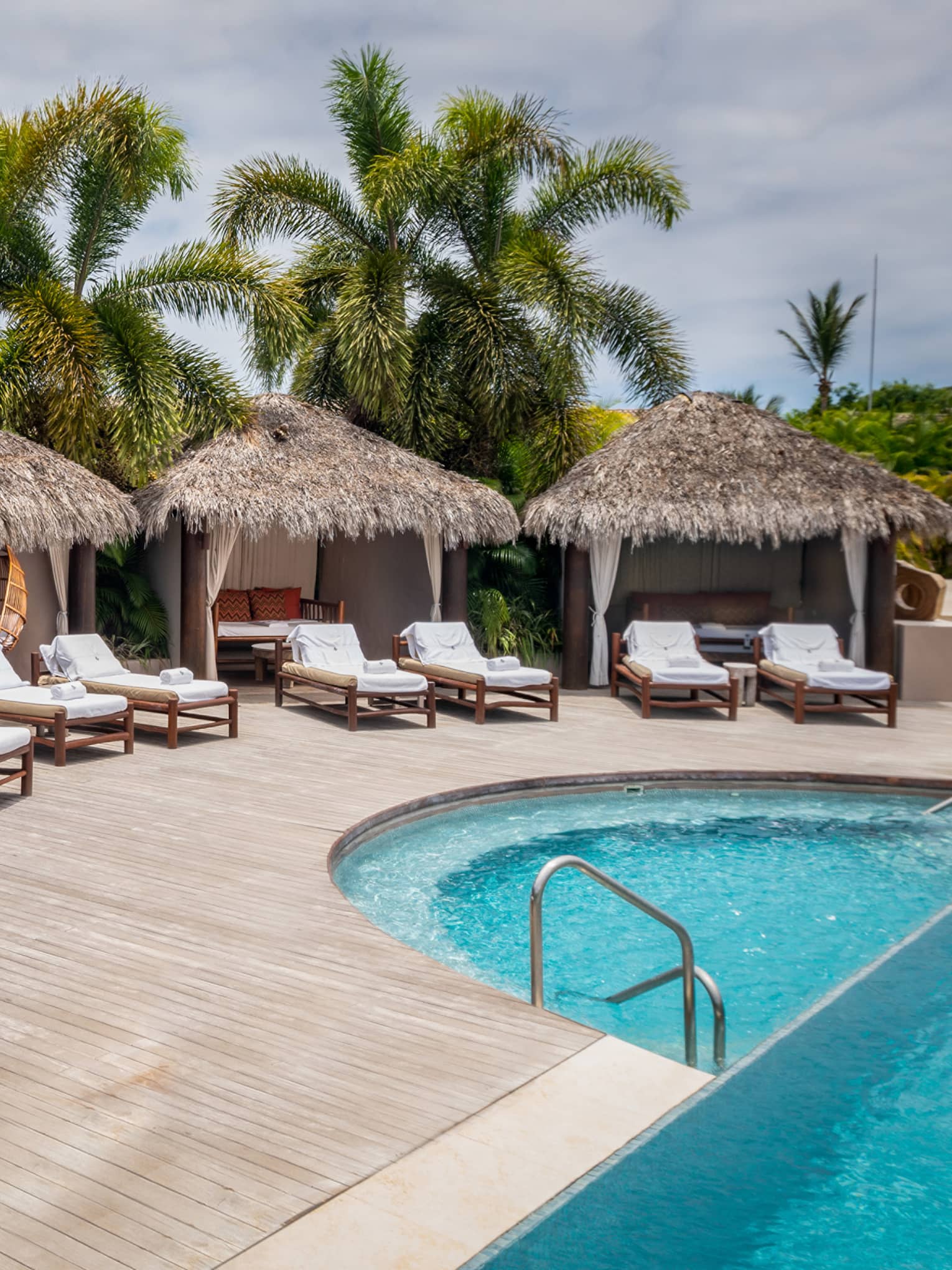 Grass-topped poolside cabanas with row of lounge chairs in front, palm trees in distance