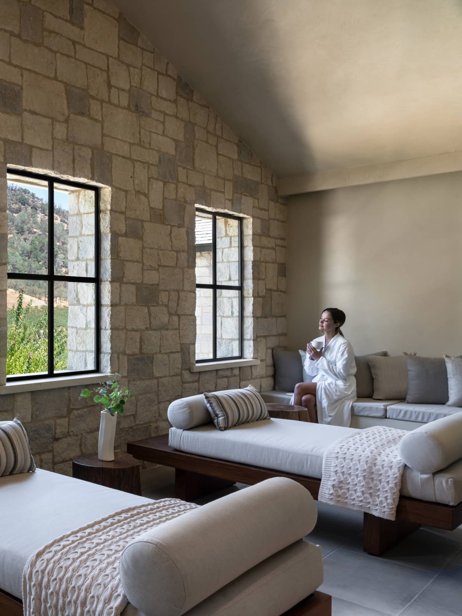 Woman in spa robe sits on couch with a tea in hand in large sunny treatment room at Spa Talisa, looking out window at Napa Valley hillside