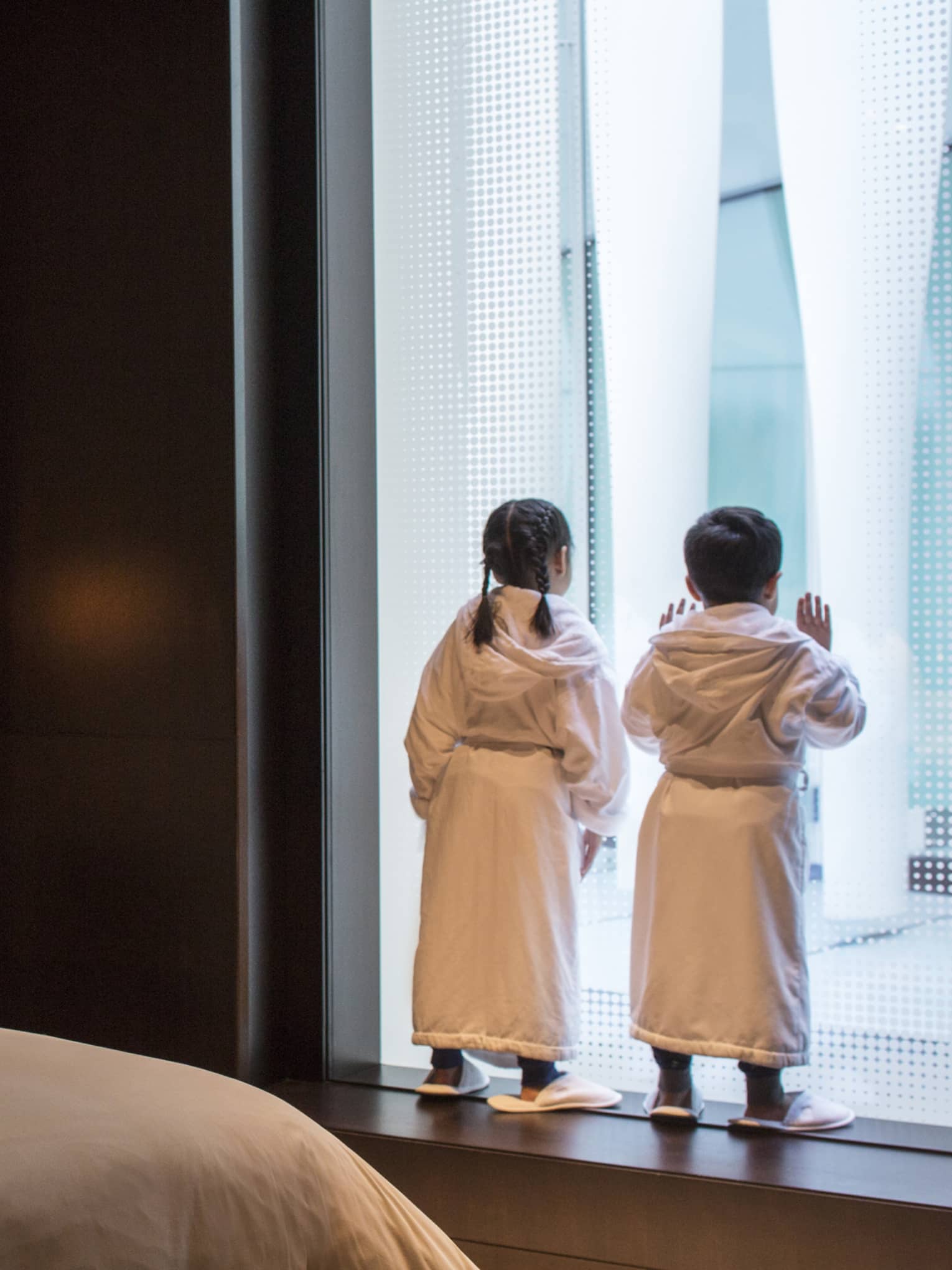 Young boy and girl in bath robes and slippers peer out the window at the city below, two teddy bears on the bed behind them