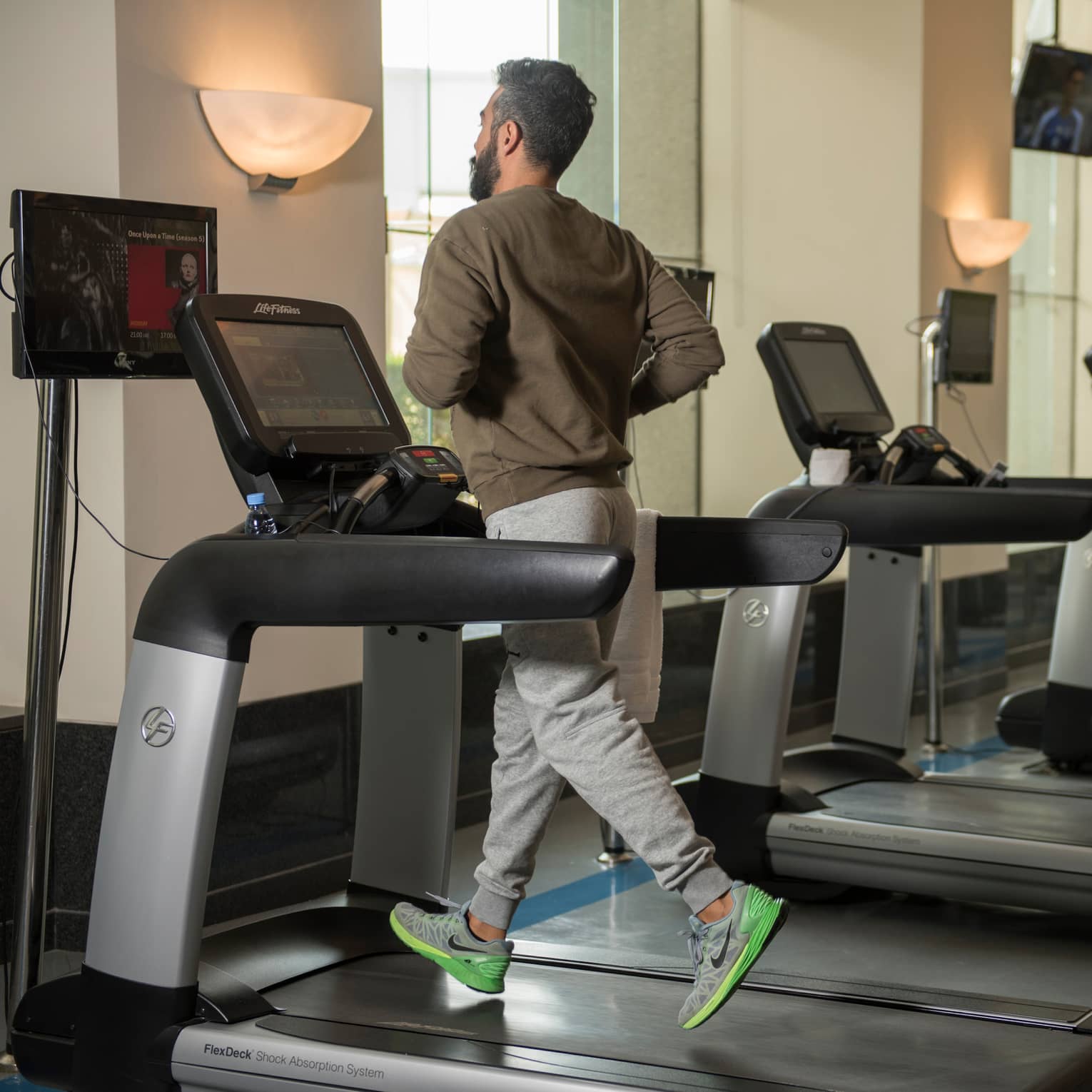 Back of man in sweatshirt and sweatpants as he runs on treadmill in gym next to row of cardio machines