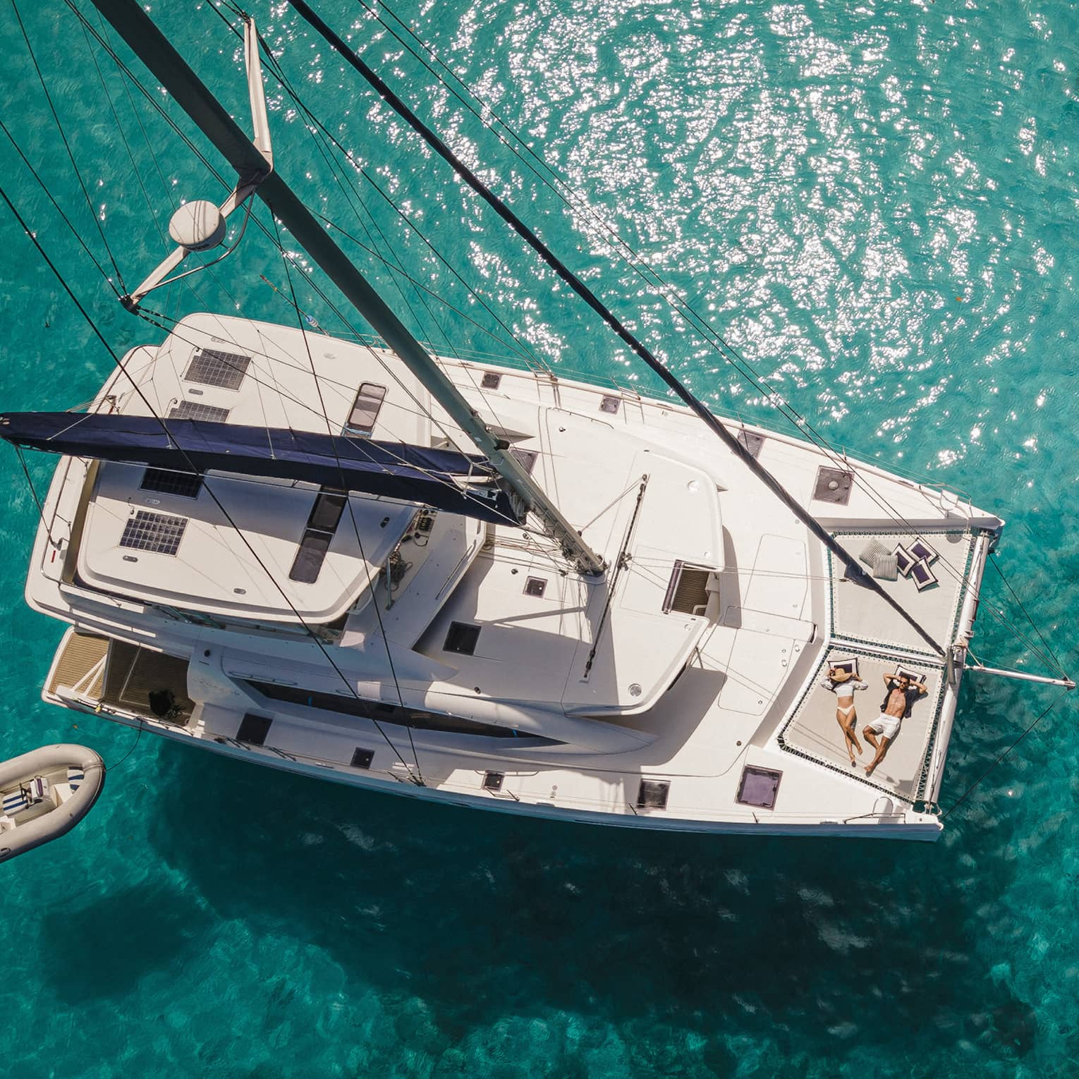 Aerial view of two sunbathers aboard a catamaran surrounded by clear, teal sun-dappled water rippling over rock formations.