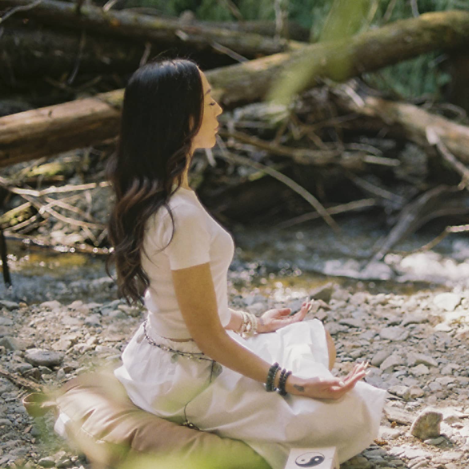 An instructor and a guest sit on yoga cushions as they peacefully meditate in a small clearing beside a brook in a forest.