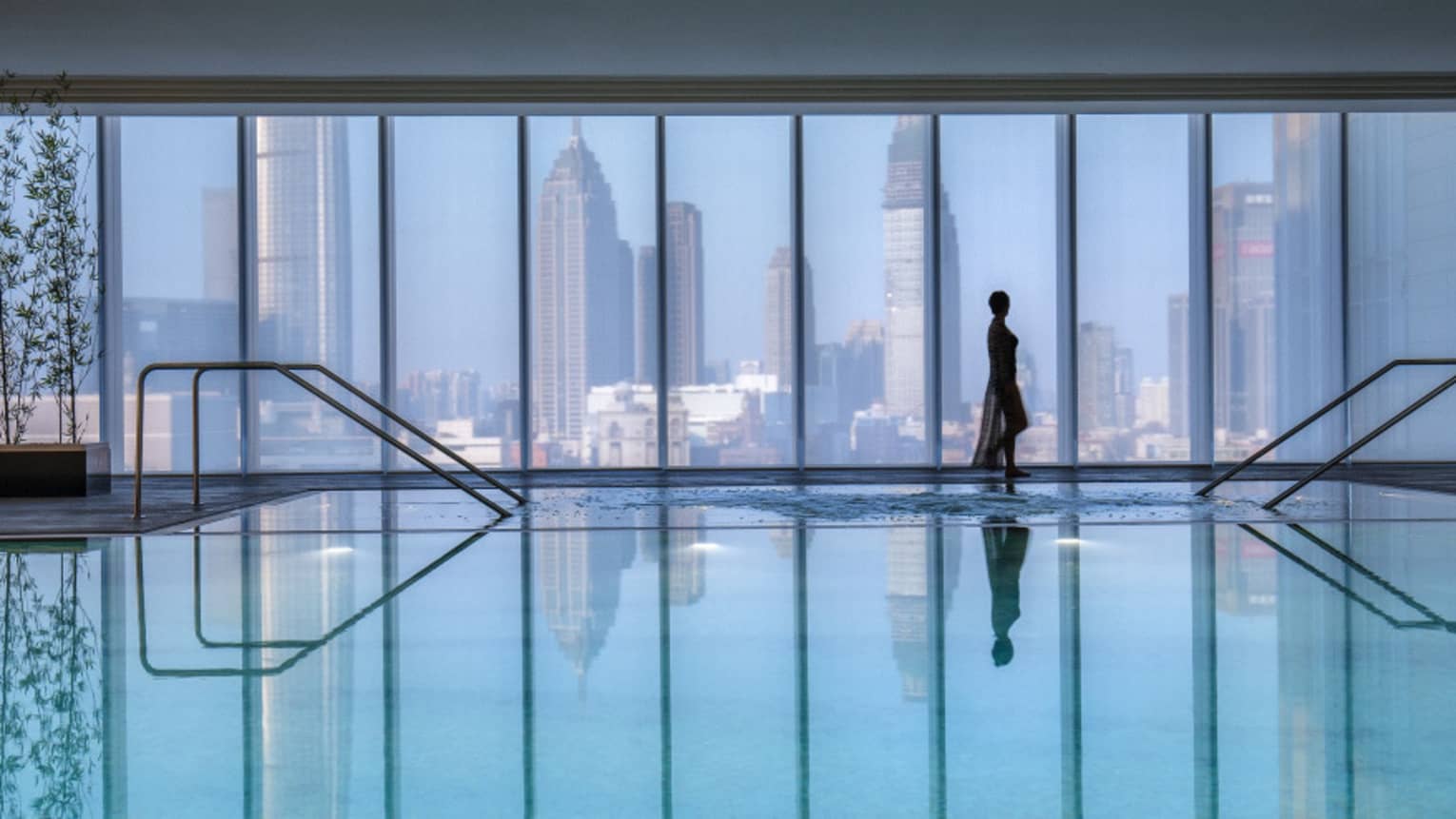 Silhouette of woman walking by blue indoor swimming pool, floor-to-ceiling window