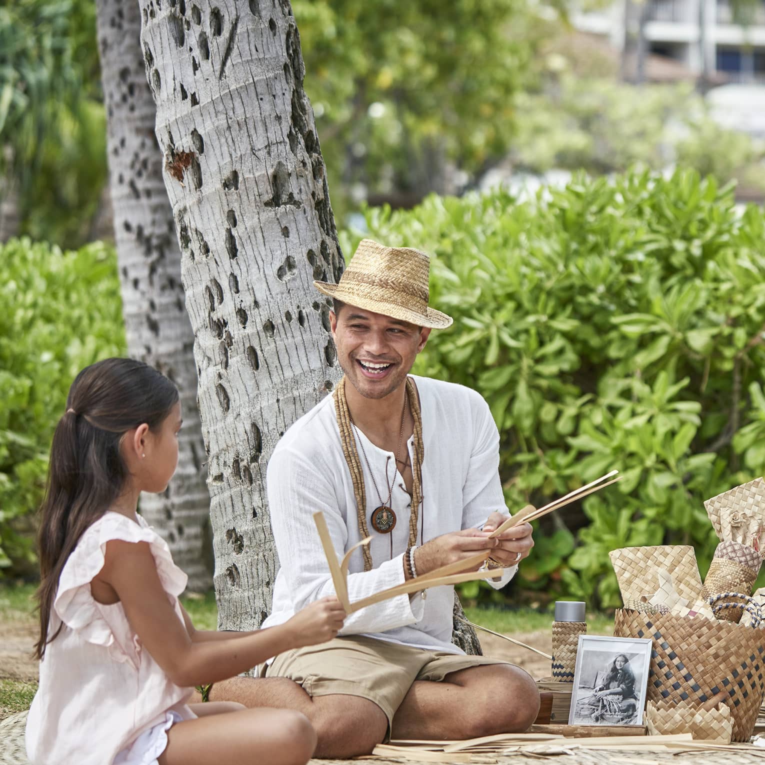 Two adults and two children sit on a blanket under a tree as they practice weaving