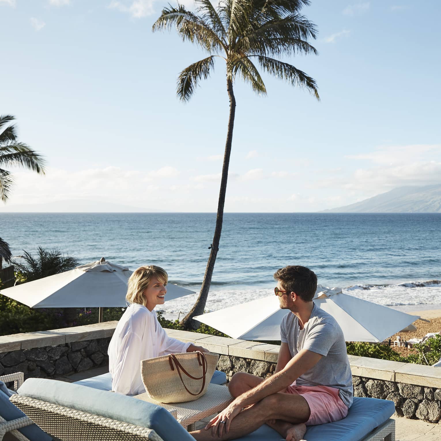 Man in shorts and smiling woman in beach shirt sit on edge of lounge chair, beach and ocean in background