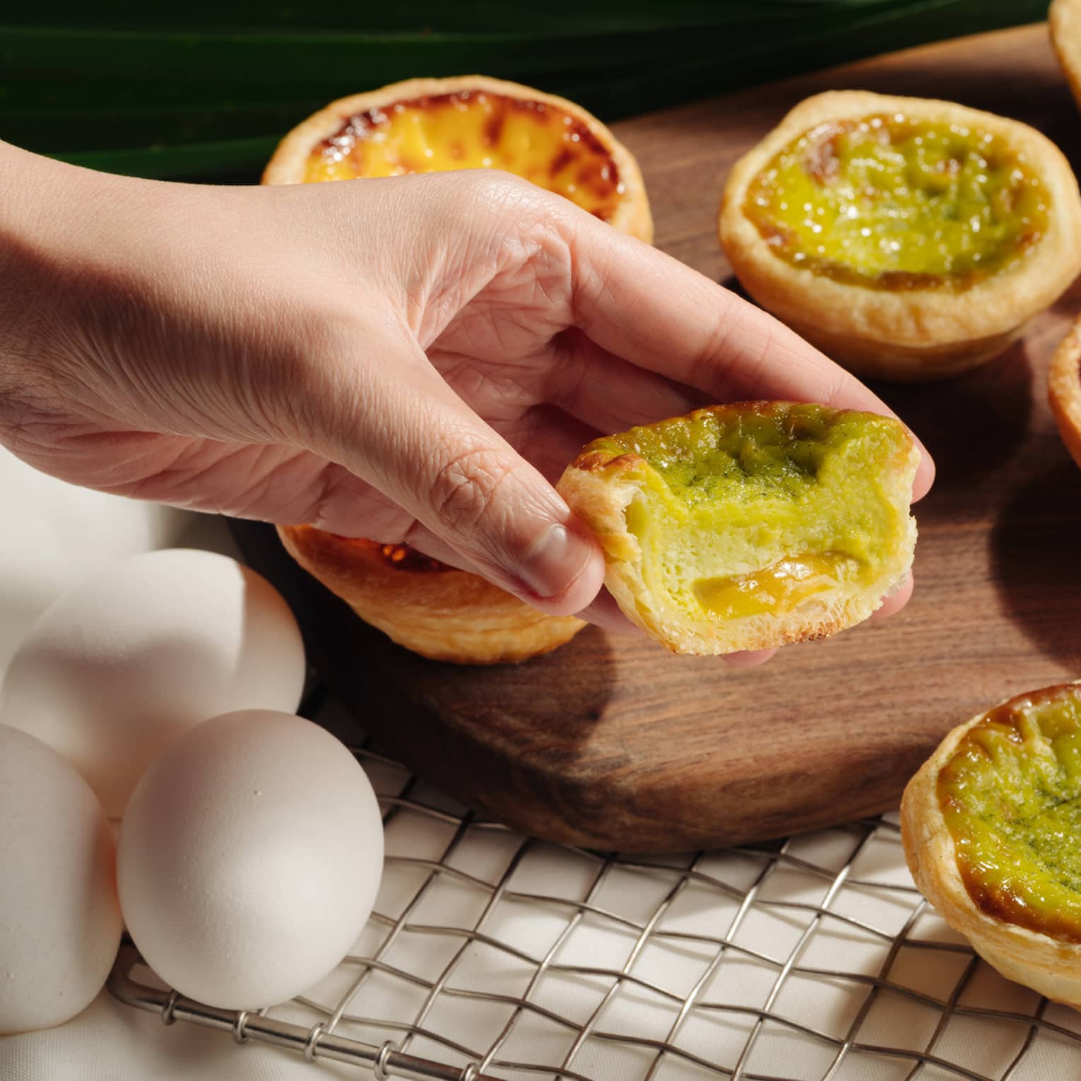 A hand holding a half-eaten Portuguese egg tart with a green filling, set against a backdrop of whole and partially-eaten egg tarts on a wooden board.