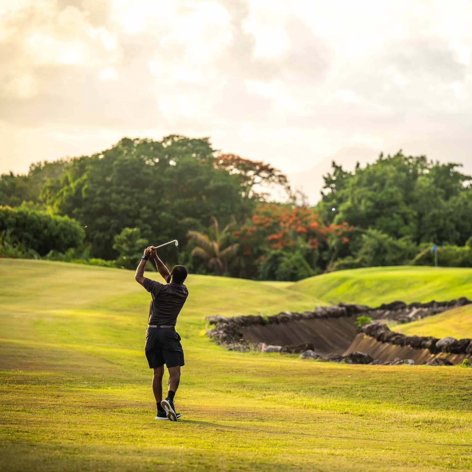 A golfer takes a swing on a bright green golf course surrounded by trees and lush vegetation, under a bright, partly cloudy sky