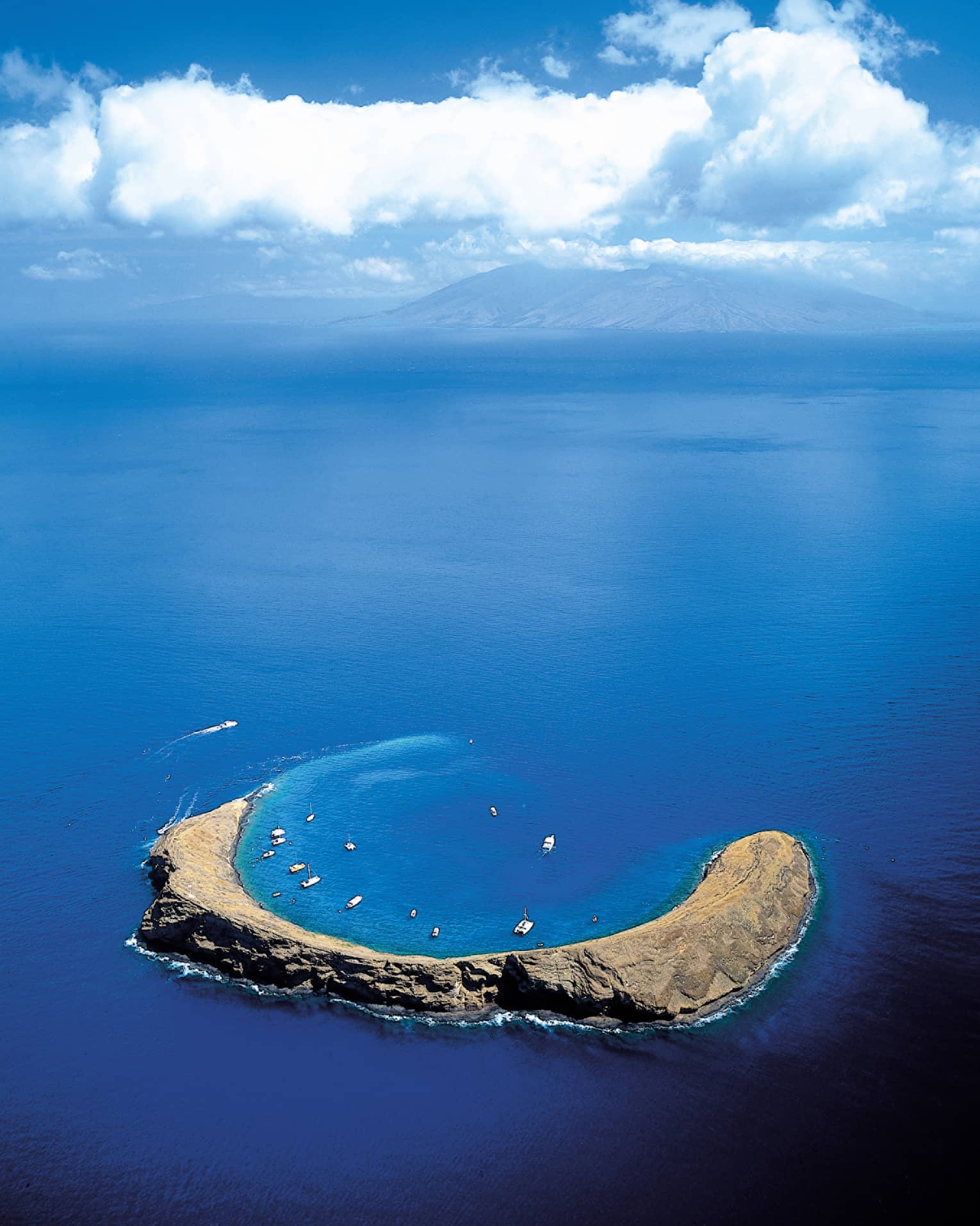 Aerial view of Molokini Crater, a crescent-shaped volcanic islet surrounded by clear blue ocean waters, with boats anchored nearby