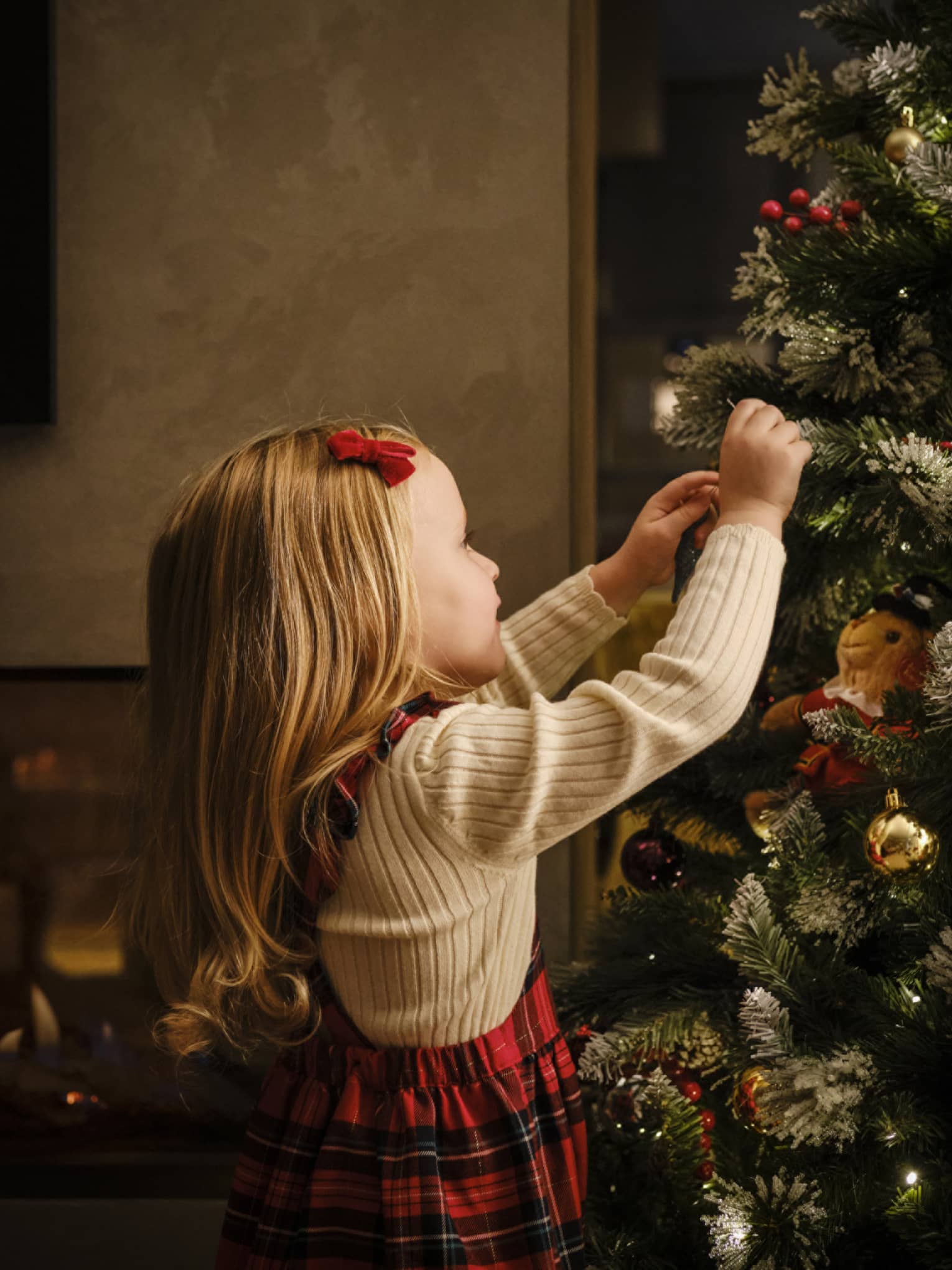 Close-up of a young child, red velvet barrette in their smooth blonde hair, arms reaching up to decorate a Christmas tree.