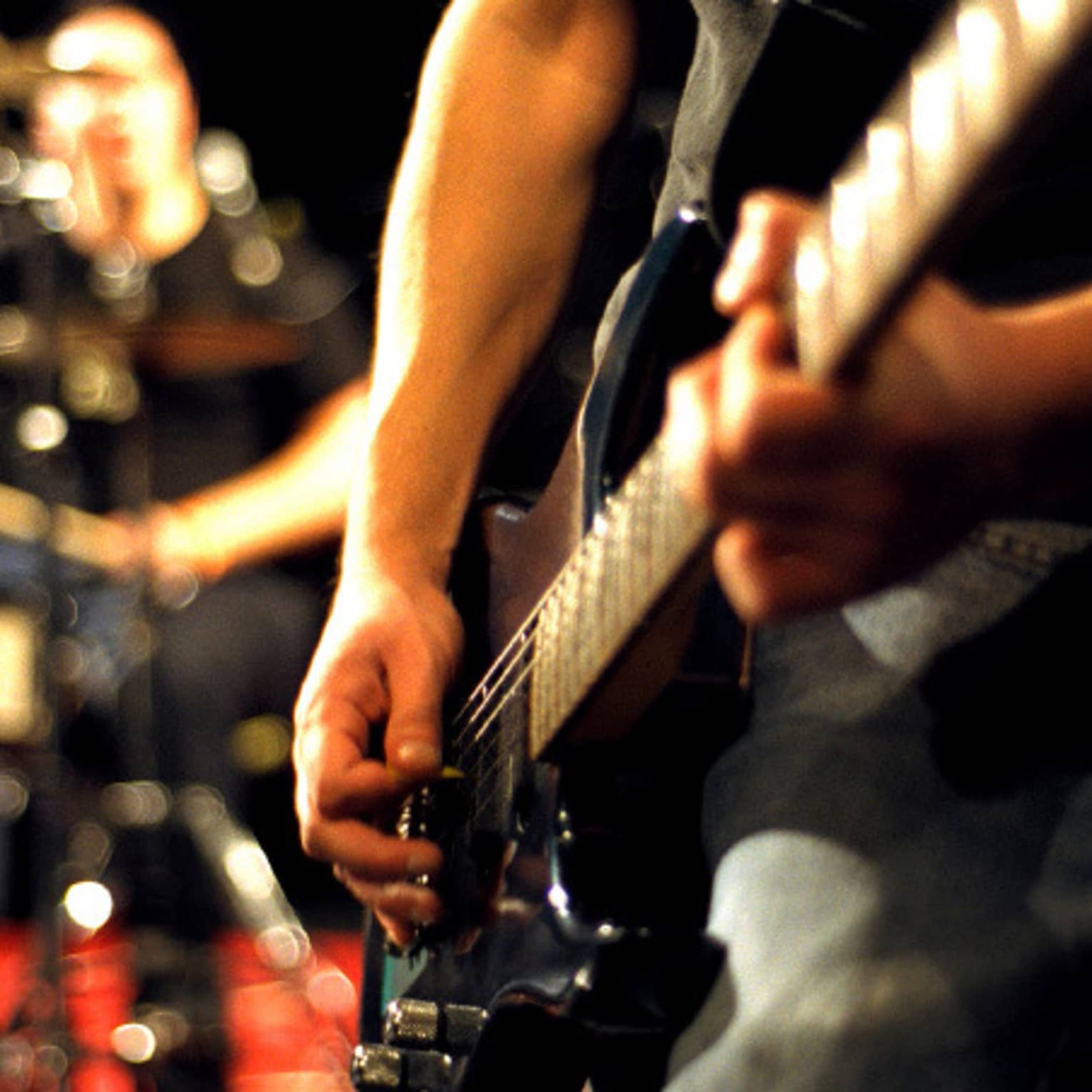 Close-up of musician's arms and hands playing an electric guitar with drumset in the background.