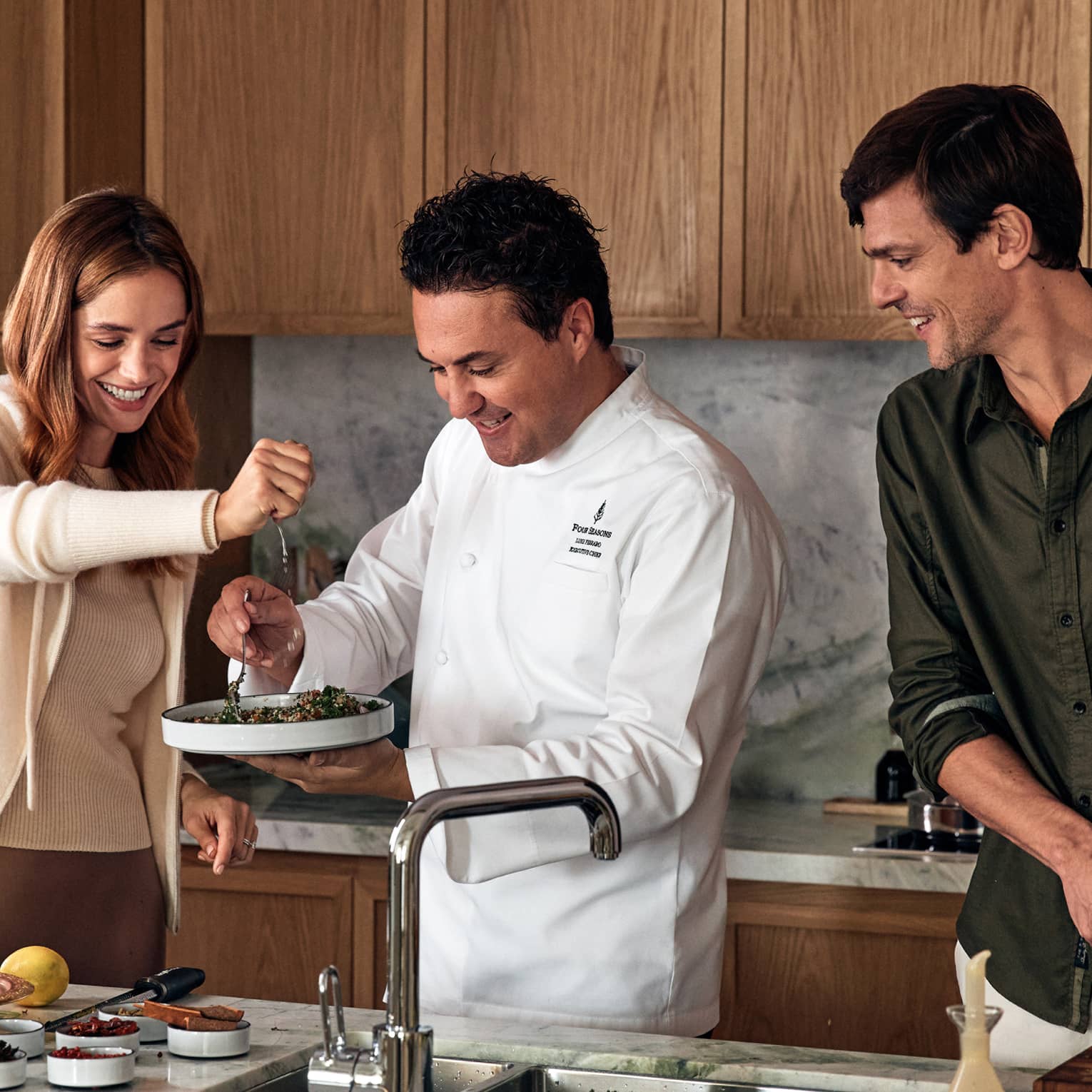 A chef holds a plate of tabbouleh as a guest squeezes a lemon overtop and another guest chops vegetables on a marble counter.