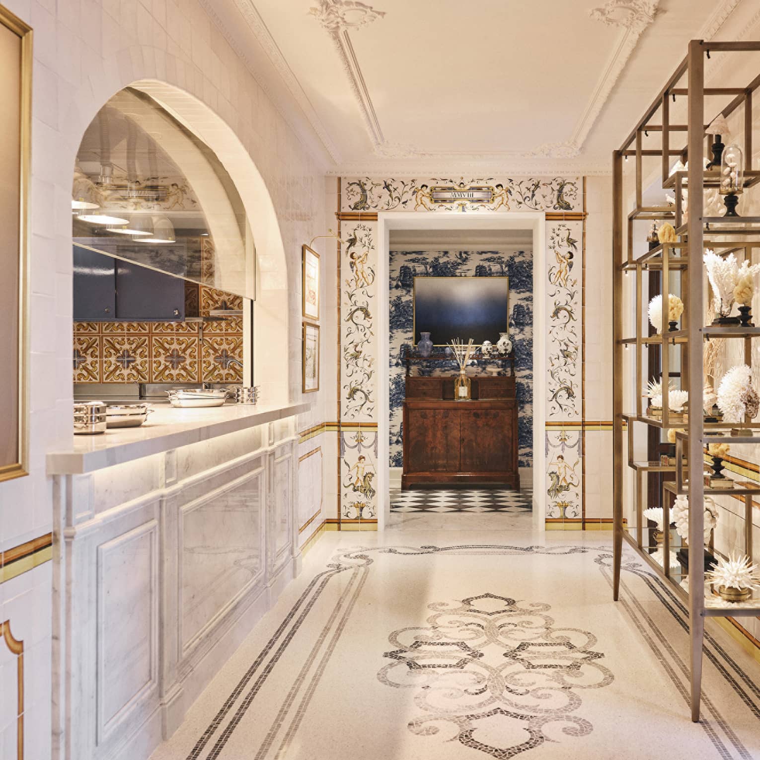 A long, bright hallway with a floor of decorative tile work. A an arched opening with a counter is along one wall and display shelves are along another wall.