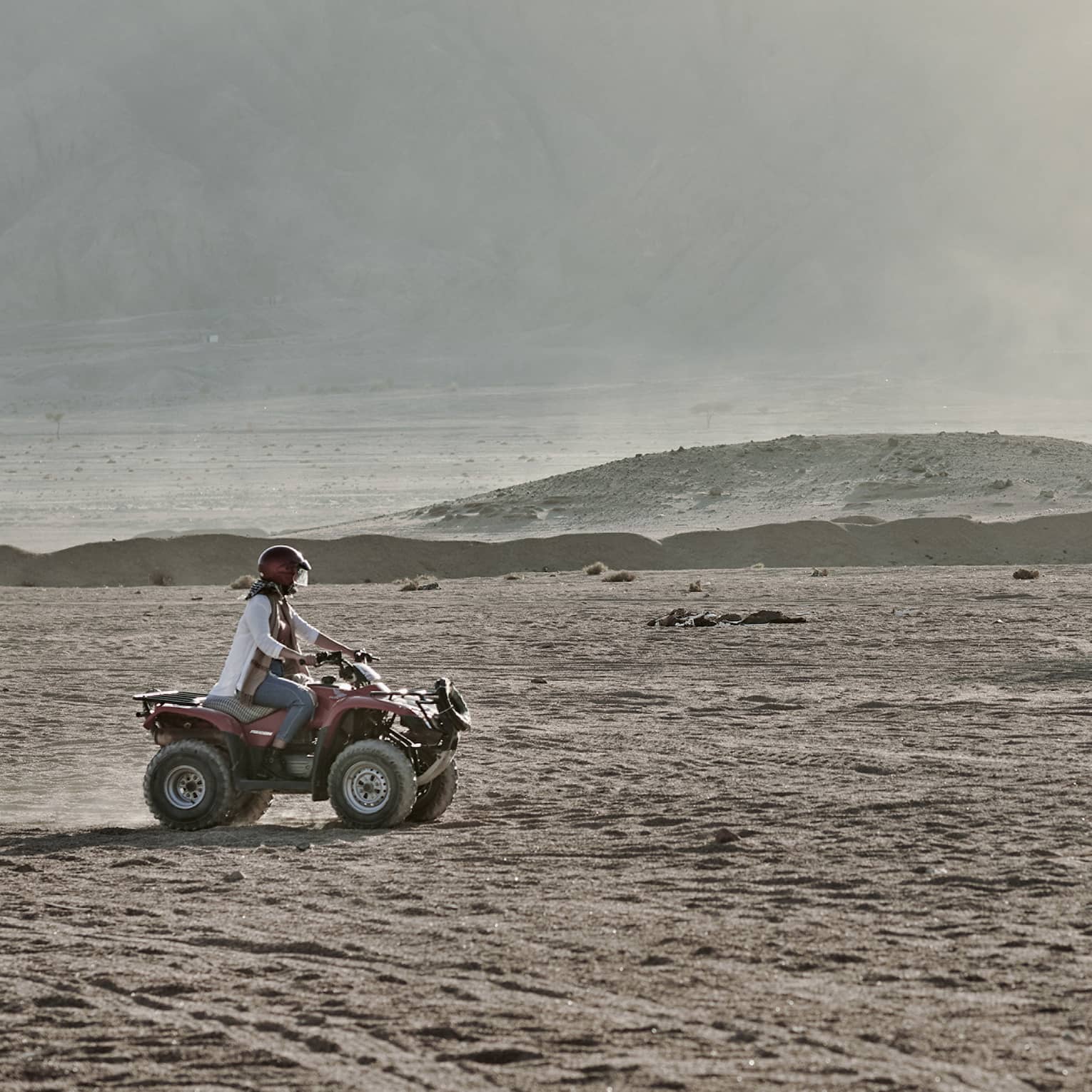 A person in a helmet rides an ATV over an arid, dusty landscape past large hazy mountains that fill most of the image.