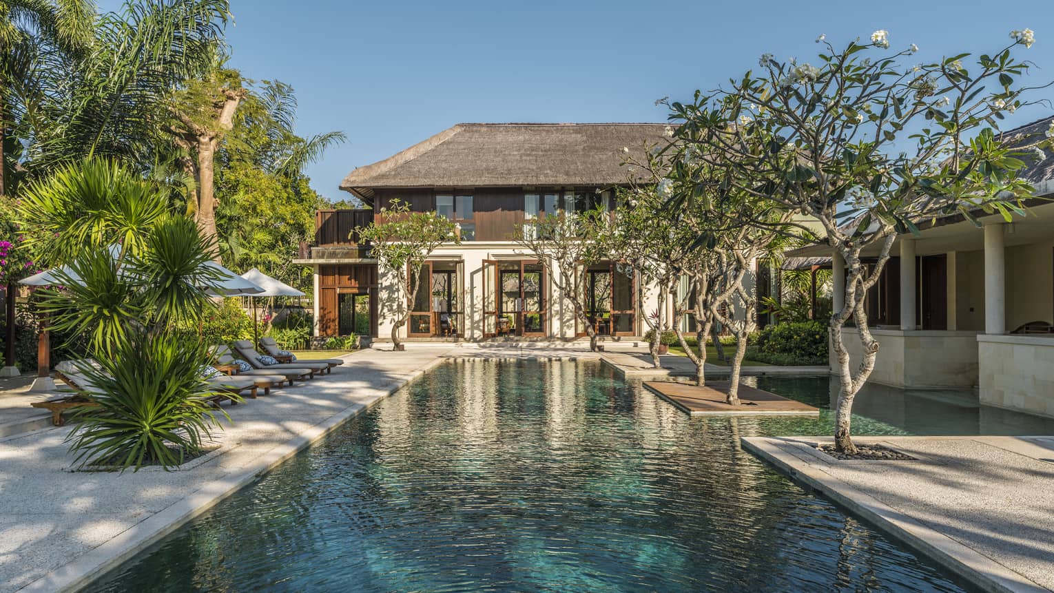 Two-storey villa at end of long outdoor swimming pool lined with trees, tropical plants